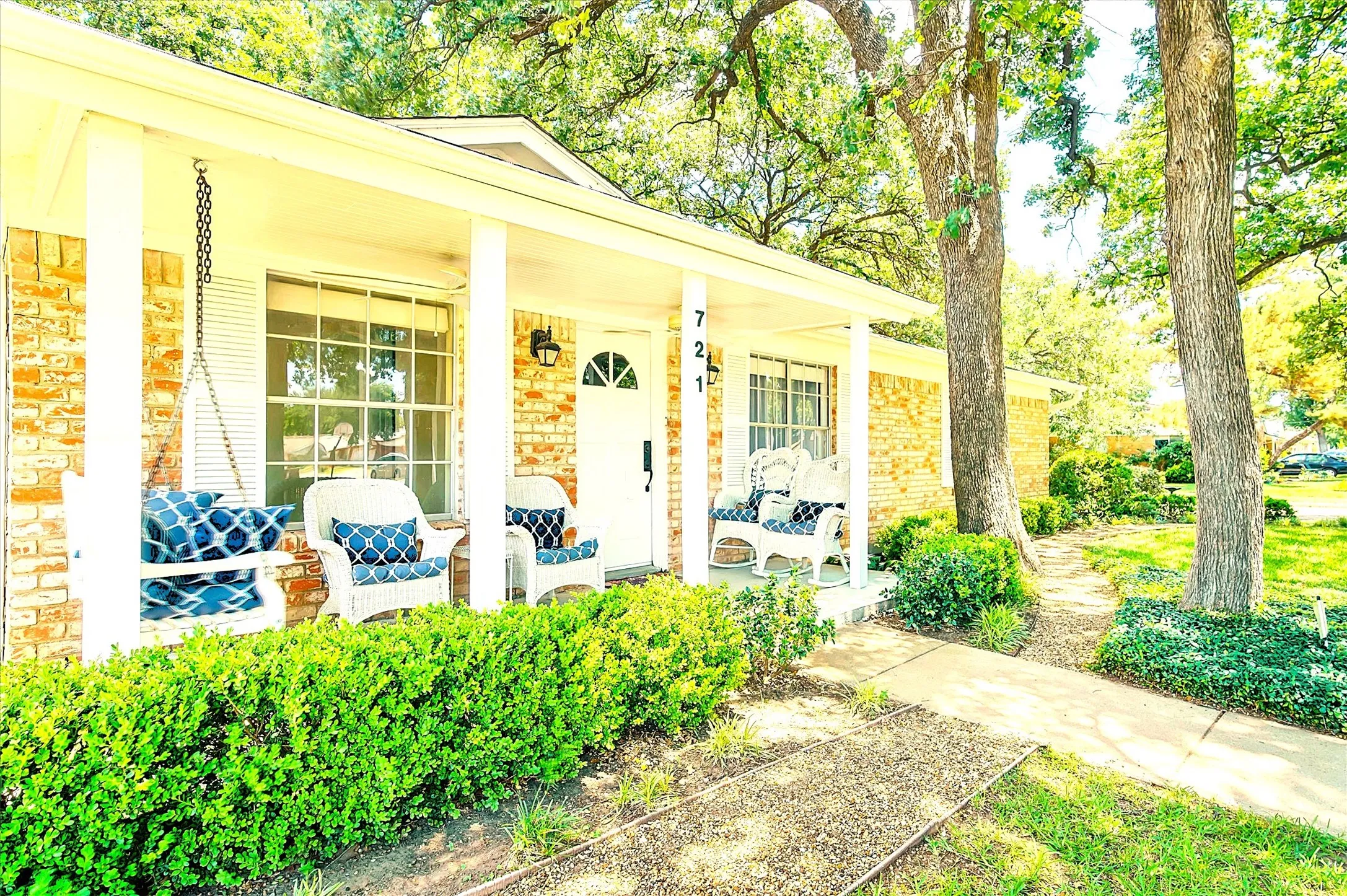 Brick exterior featuring a covered porch with white columns, white front door with an arched window, and a pathway bordered by mature trees and manicured shrubs