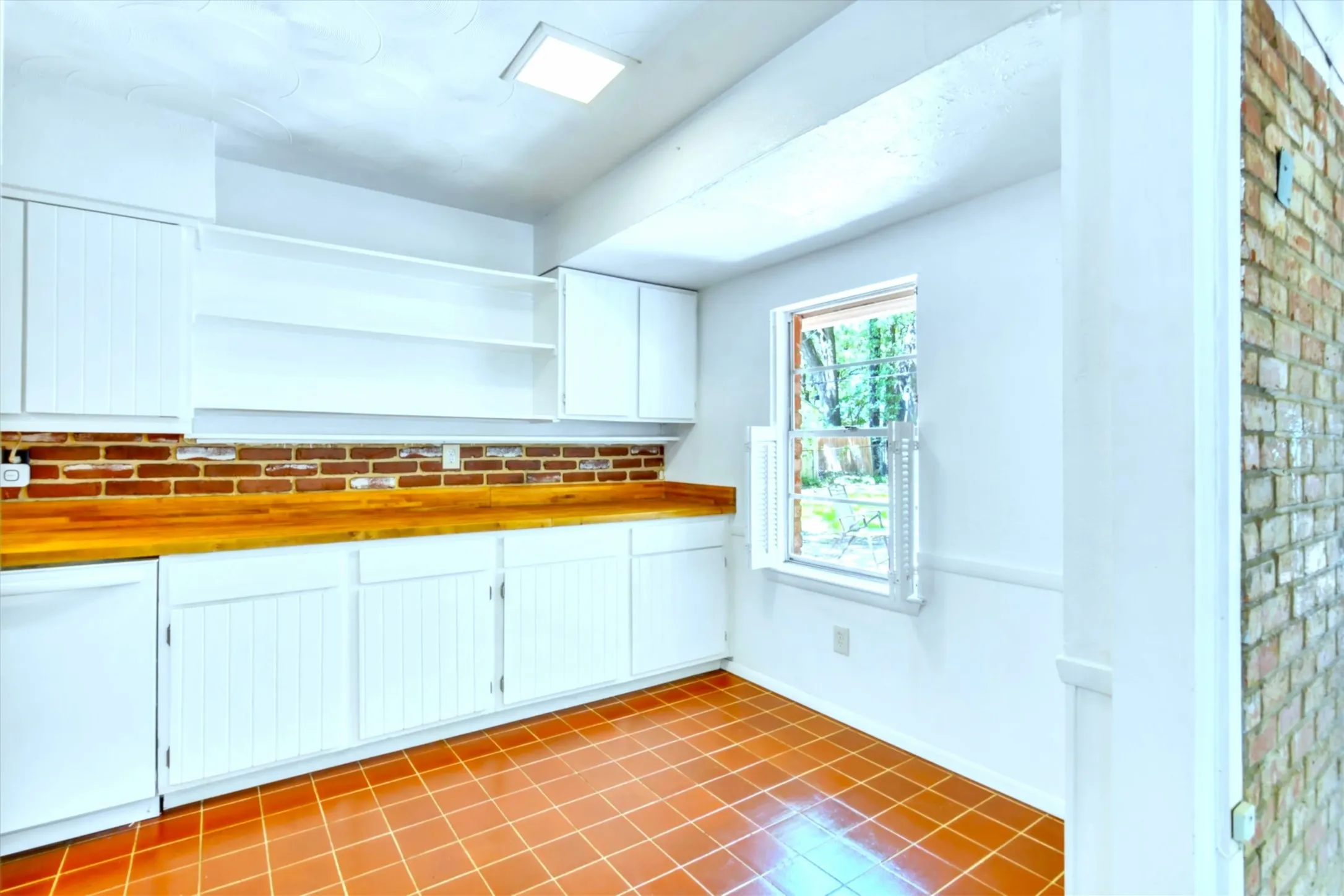 Galley kitchen featuring terracotta tile flooring, white shaker cabinetry with beadboard accents, wood countertops, and a brick backsplash
