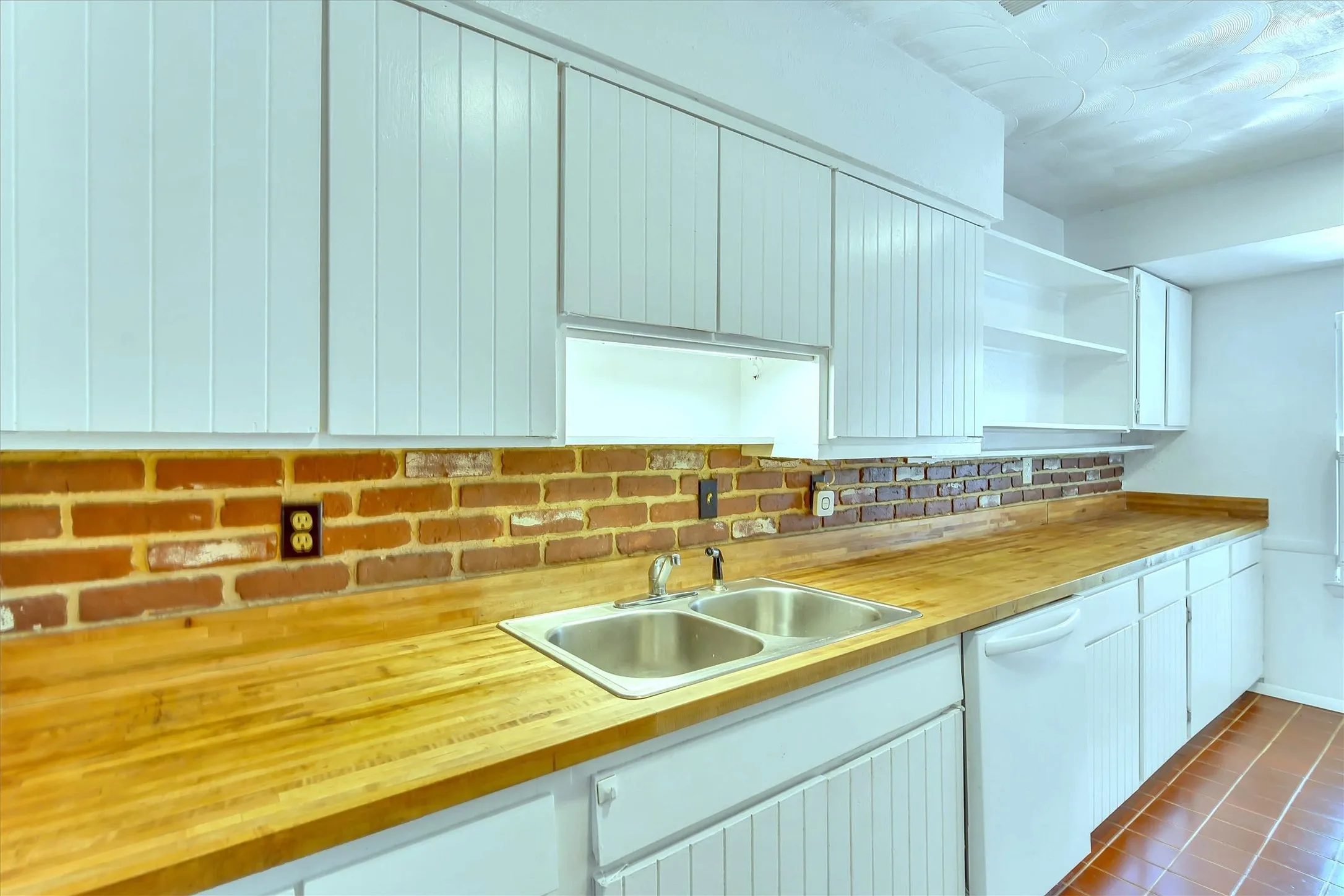 Kitchen featuring white cabinetry, wood-finish countertops, a stainless steel double-basin sink, brick-style backsplash, and a built-in dishwasher