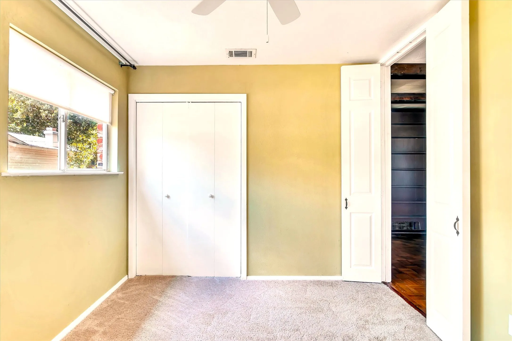 Carpeted room featuring a window with a roller shade, a white bi-fold closet, and a ceiling fan