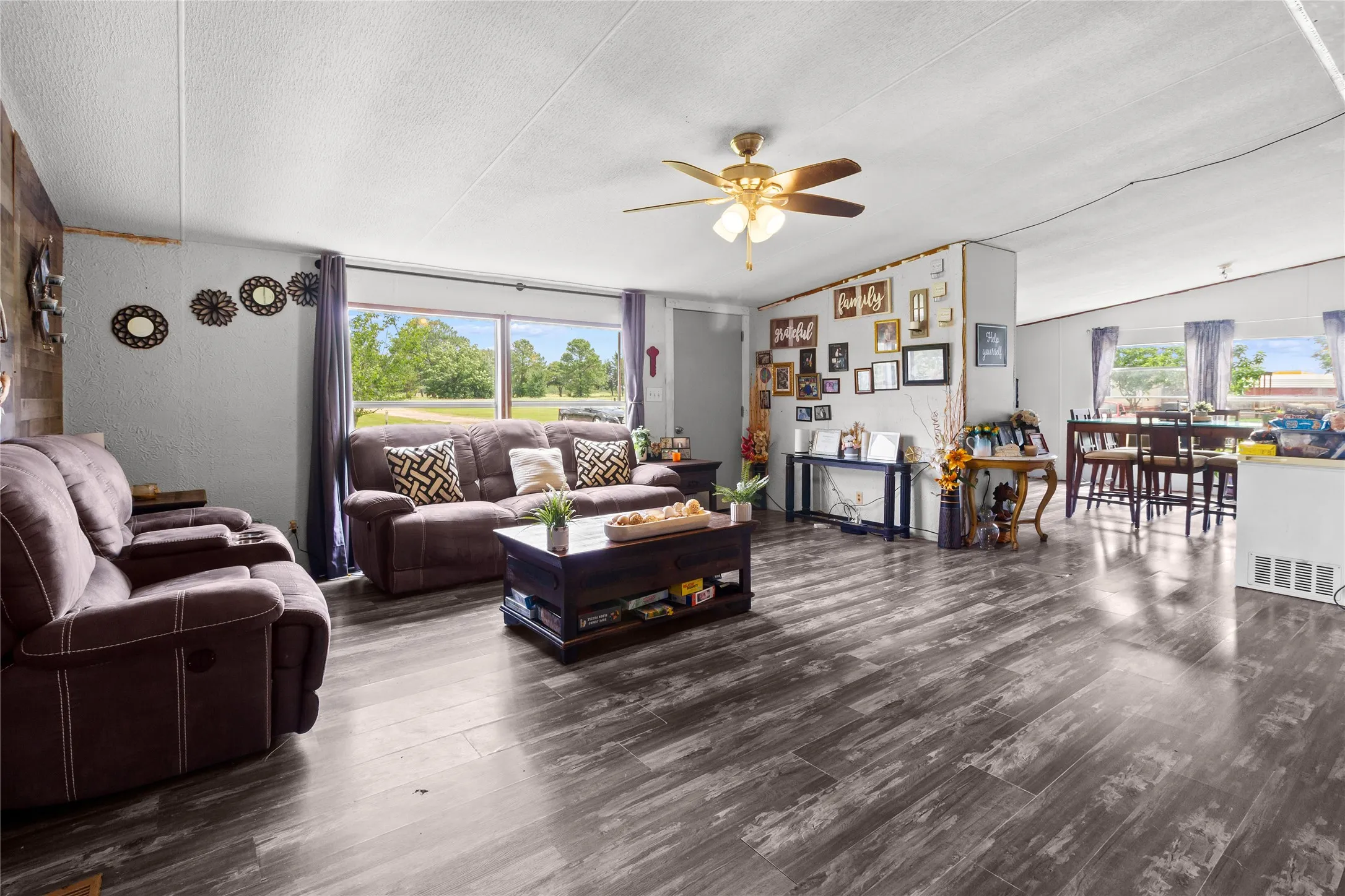 Spacious living area featuring wood-finish flooring, a ceiling fan with integrated lighting, and multiple windows