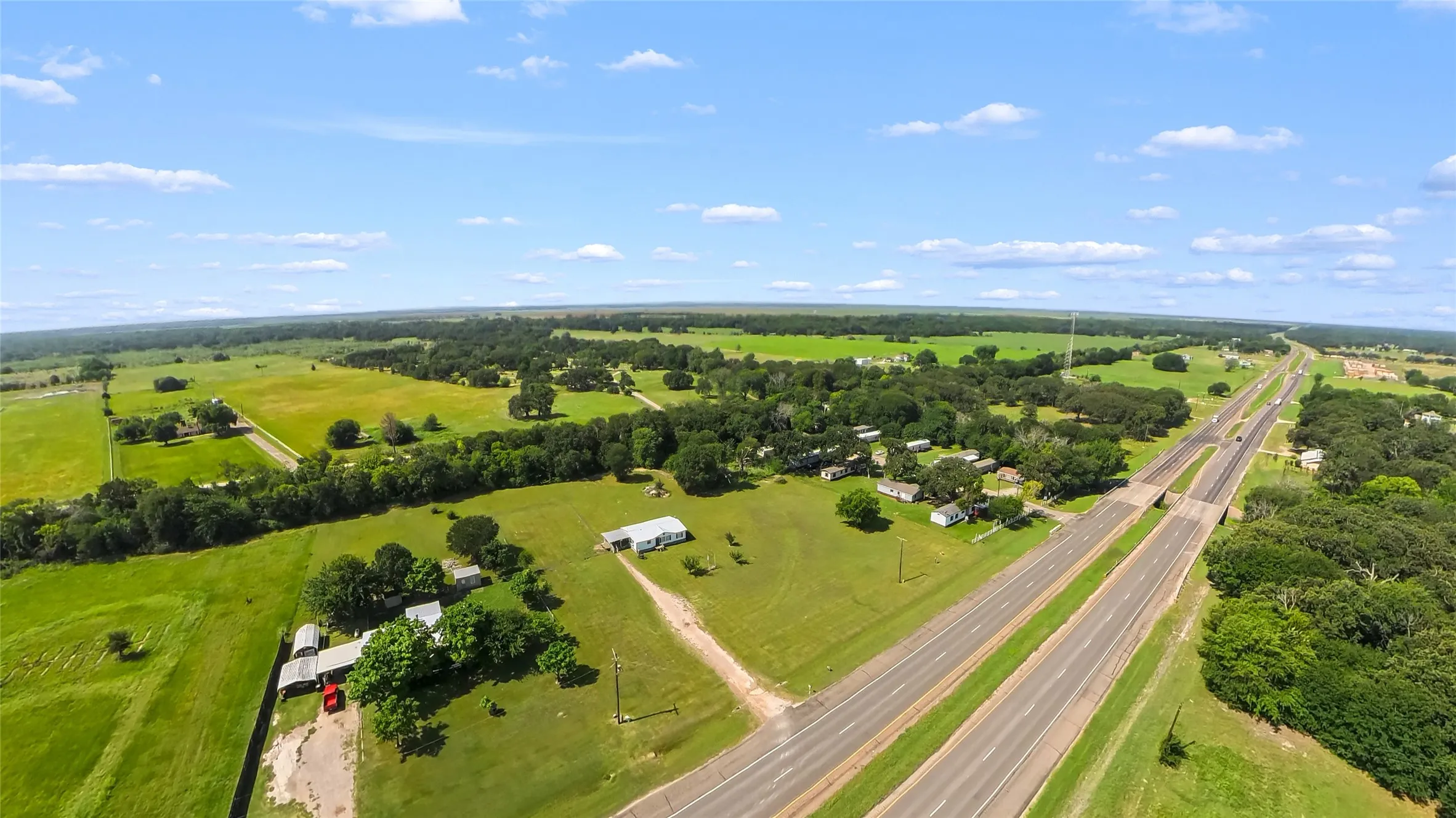 Expansive rural landscape featuring extensive green fields, mature trees, a multi-lane highway, and scattered residential structures