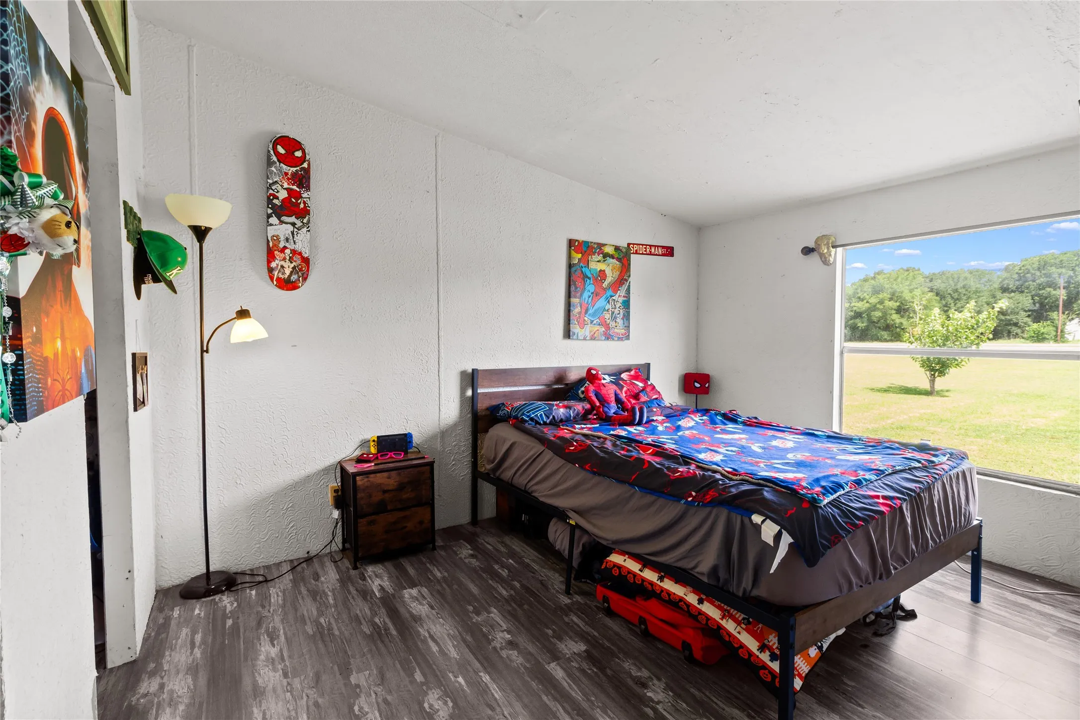 Bedroom with wood-finish flooring, a broad window overlooking outdoor greenery, and a white textured wall finish