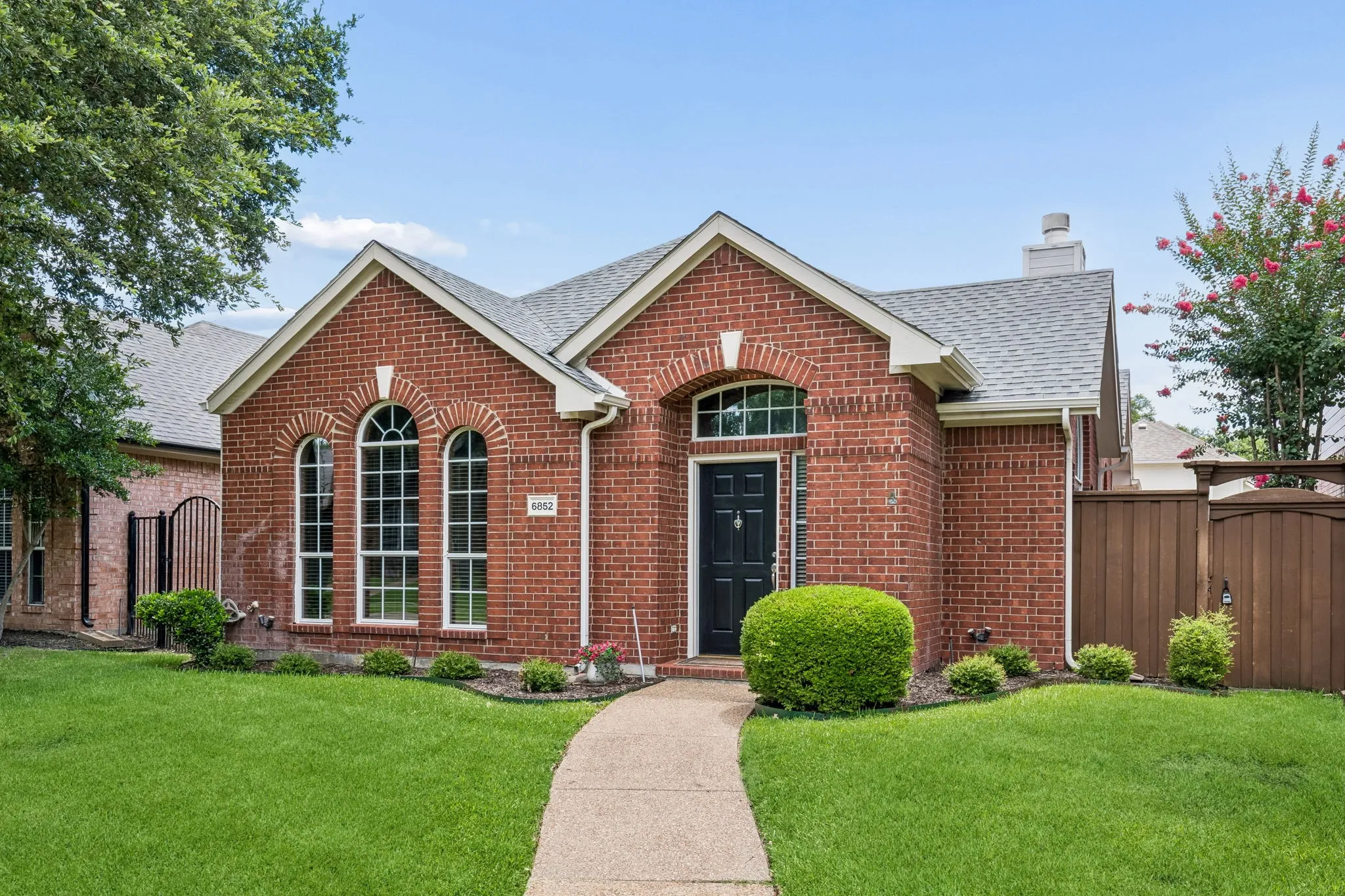 Red brick facade featuring a dark entry door with transom window, arched windows, a concrete pathway, a shingle roof, and a wood privacy fence