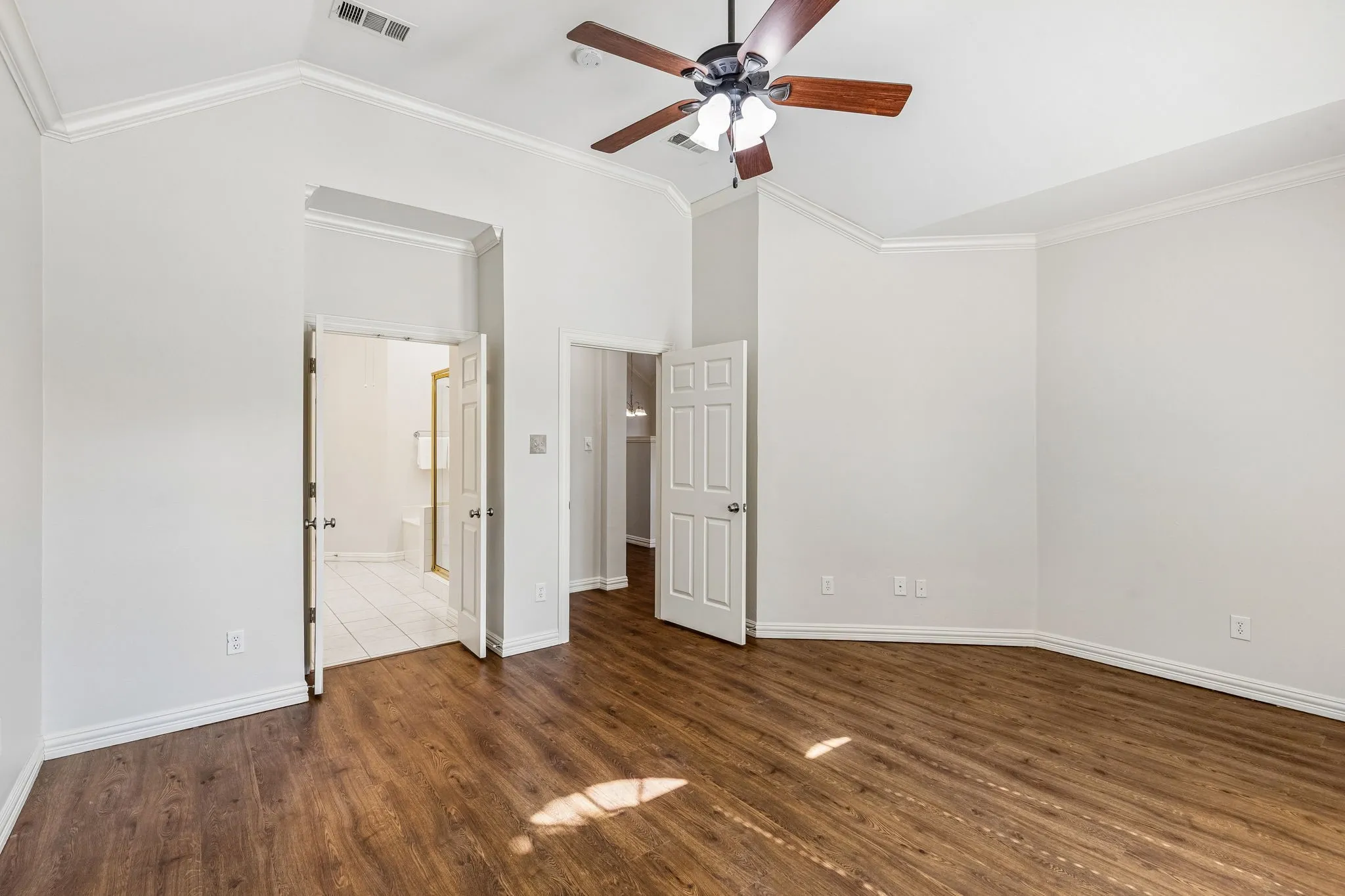 Spacious bedroom featuring rich wood-finish flooring, ceiling fan with integrated lighting, white crown molding, and multiple closet entries