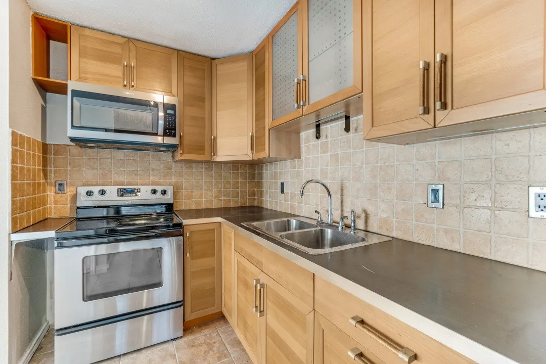 Kitchen featuring natural wood-finish cabinetry with modern hardware, stainless steel appliances including an oven range and microwave, a double basin sink, and a tiled backsplash