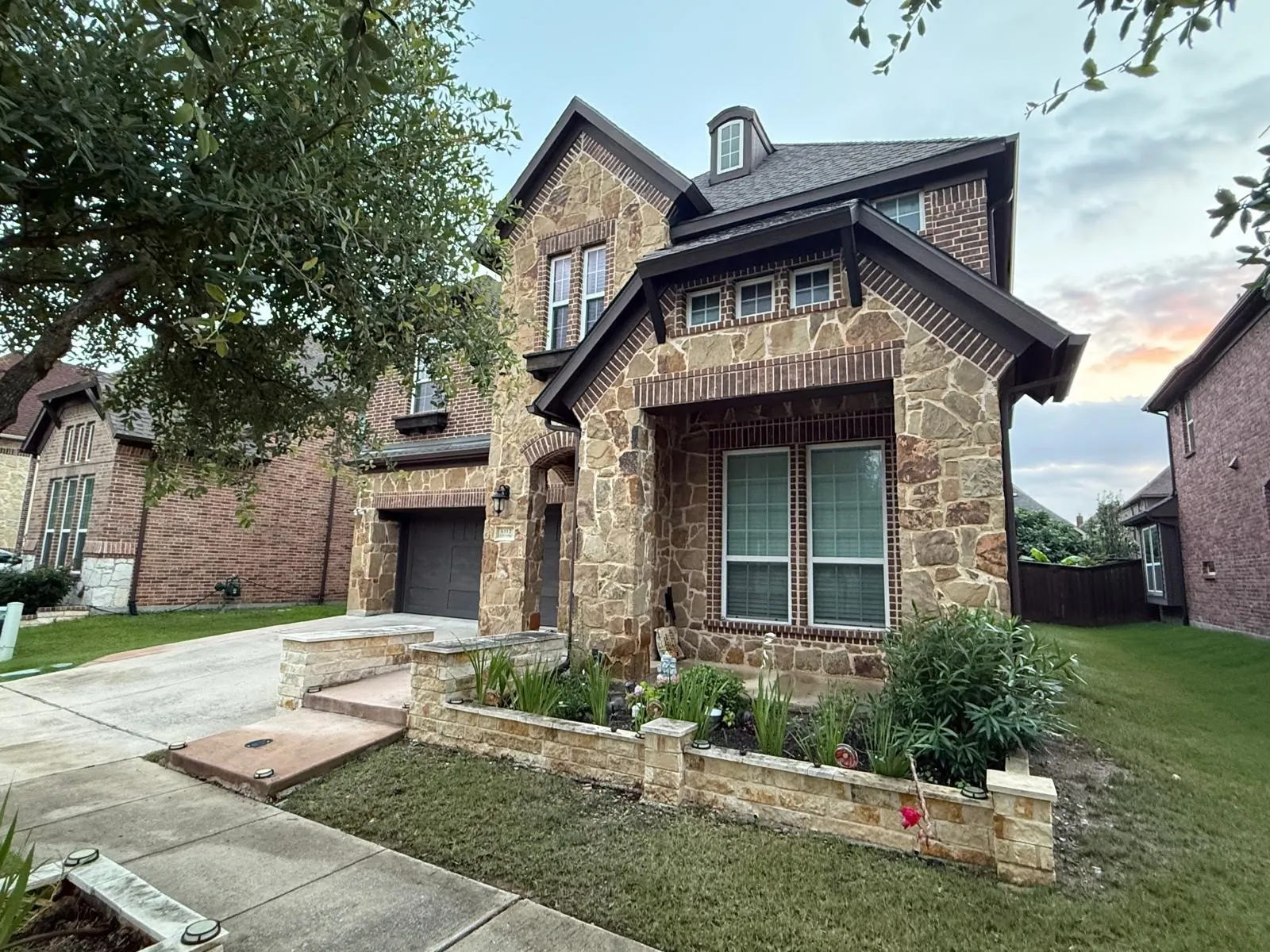 Two-story residence featuring a stone and brick facade, an attached garage, a dormer window, and a planted stone planter box