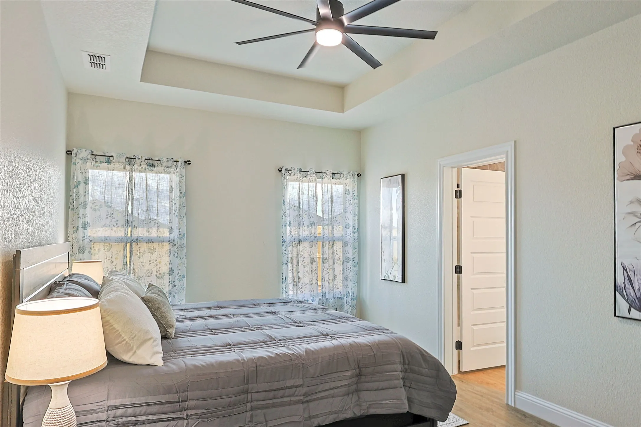 Room featuring a tray ceiling with a ceiling fan, two windows with sheer curtains, a five-panel interior door, and wood-finish flooring