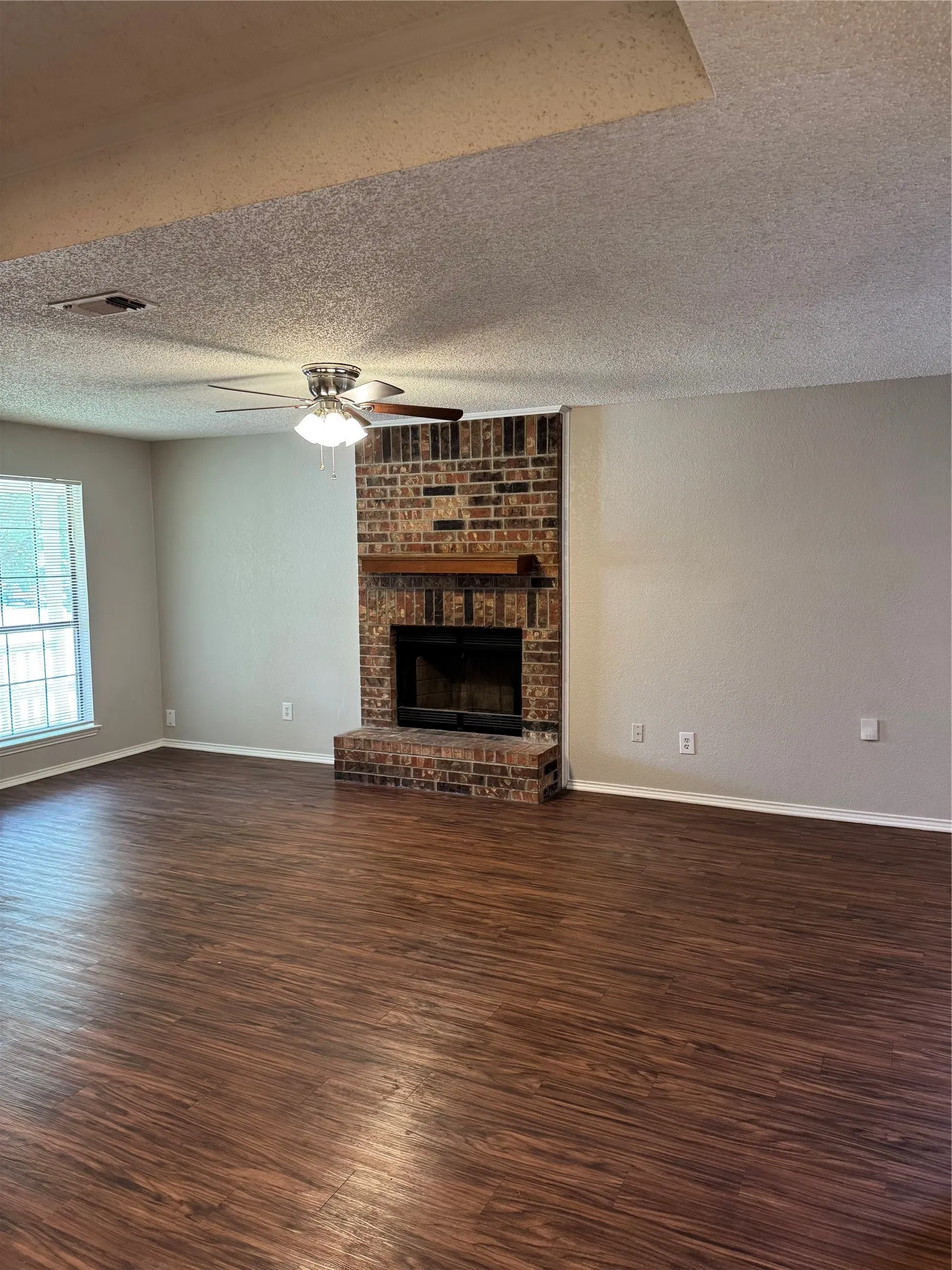 Expansive room featuring wood-finish flooring, a brick fireplace with a wood mantle, a ceiling fan with light fixture, and a window with white blinds