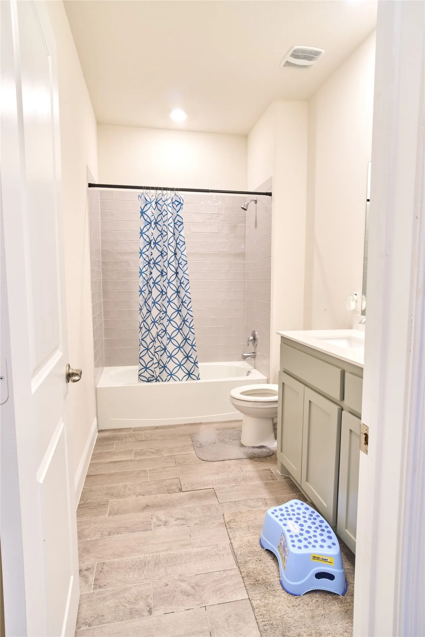 Full bathroom featuring a white tub with subway tile surround, single vanity with light gray cabinetry, and wood-finish tile flooring