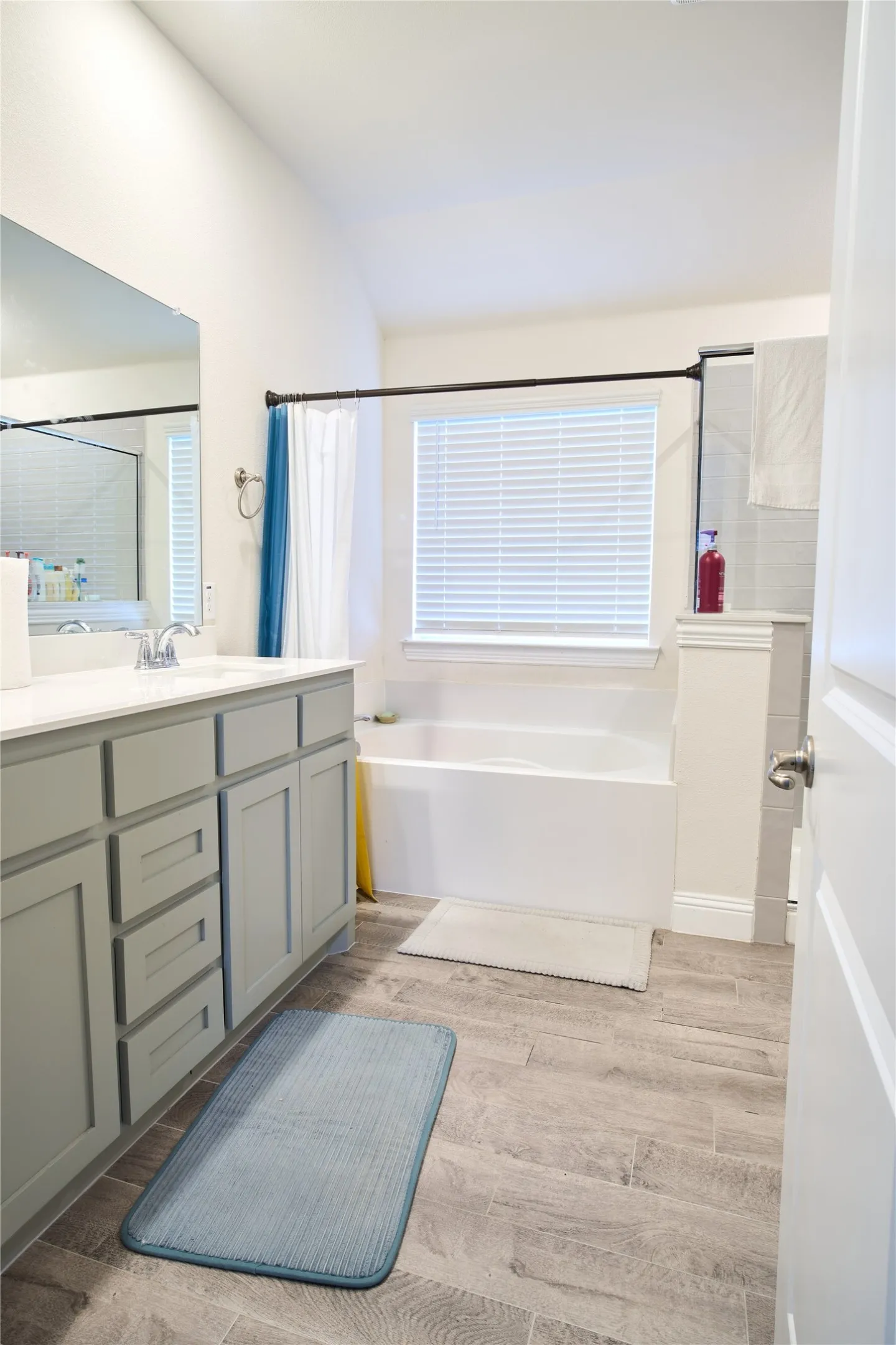 Bathroom featuring wood-finish flooring, a double vanity with grey cabinetry and white countertops, a built-in bathtub, and a window with blinds