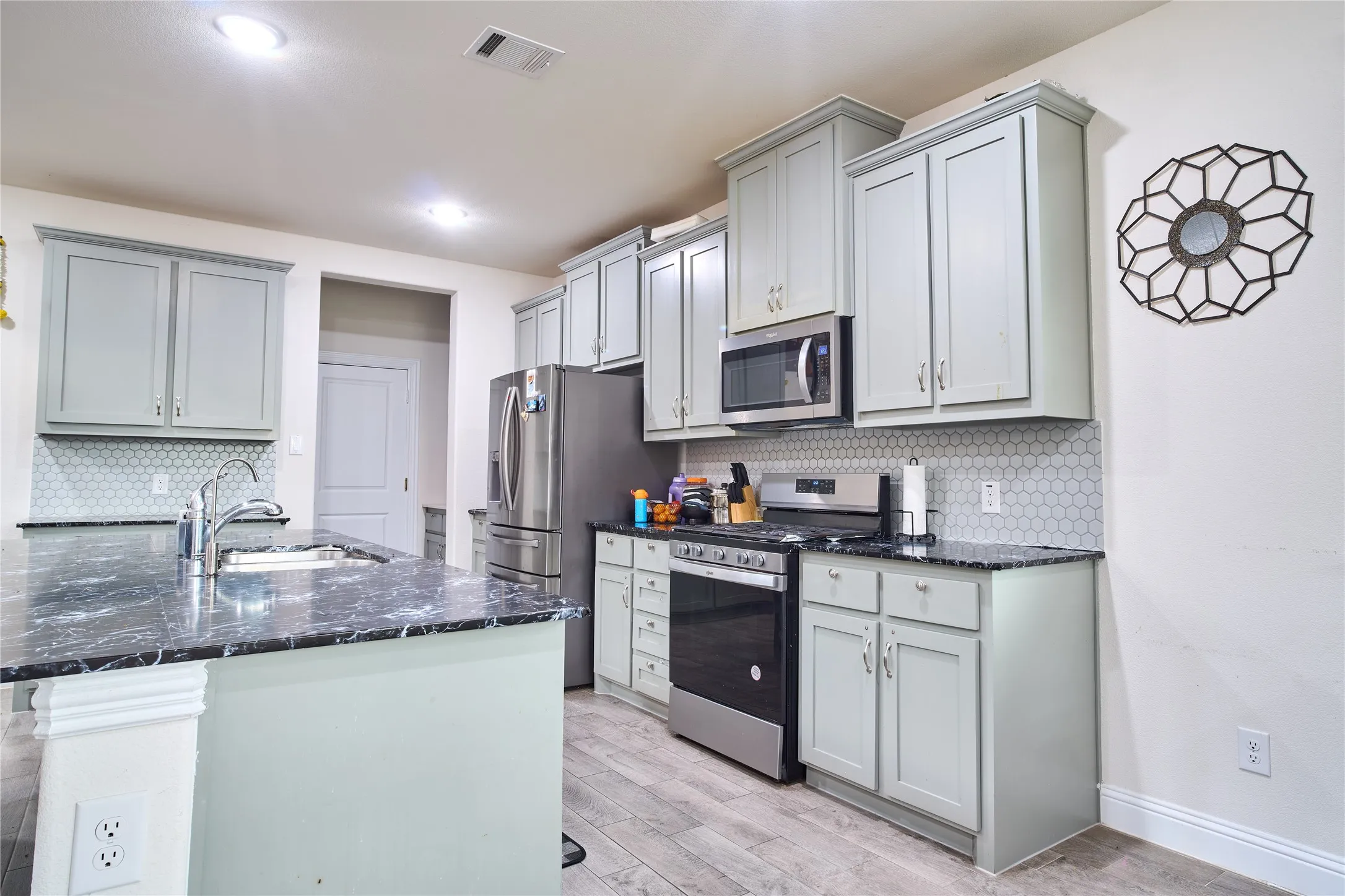 Kitchen featuring a central island with a sink, light grey cabinetry, stainless steel appliances, dark countertops, and a white hexagonal tile backsplash
