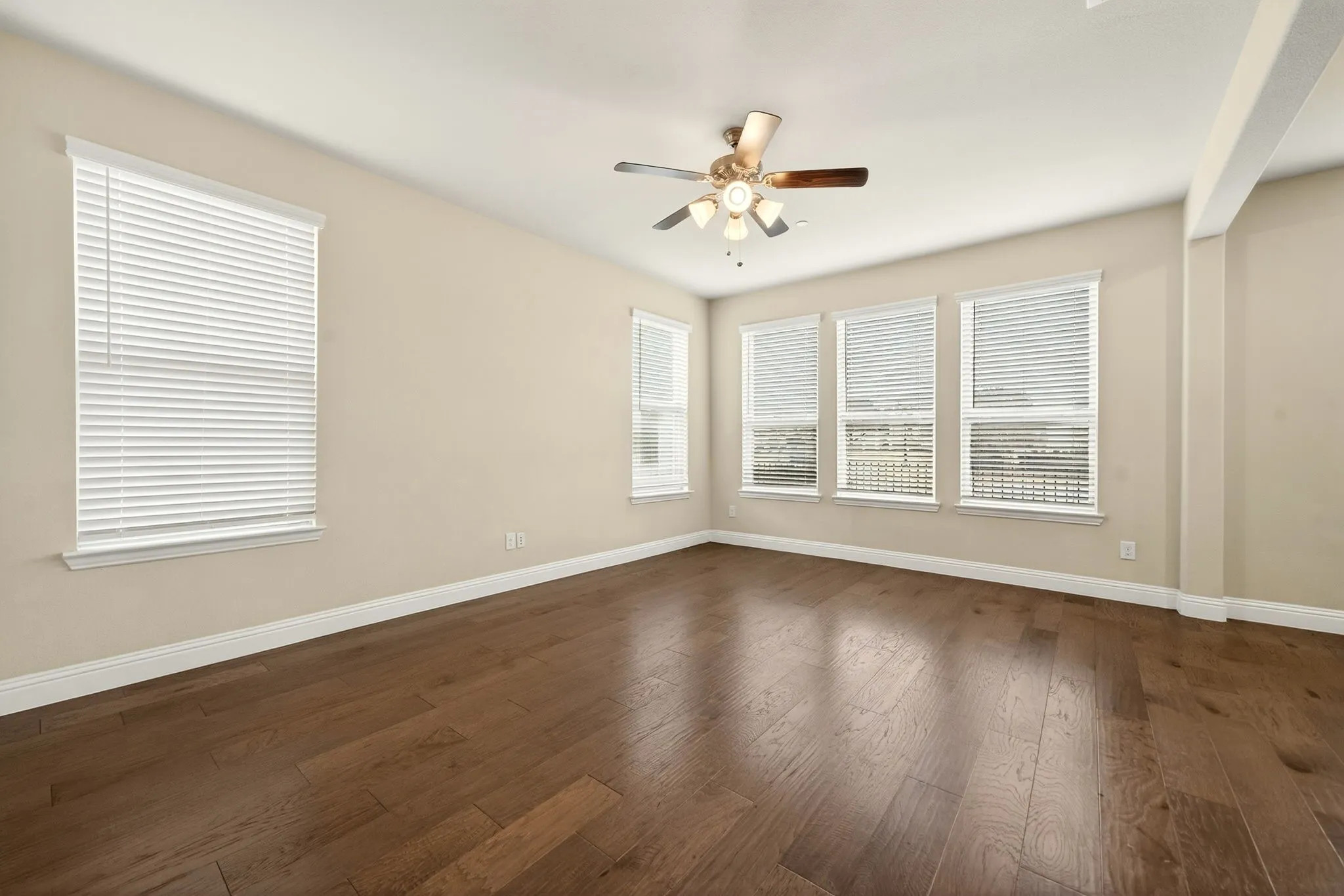Expansive room featuring rich wood-finish flooring, multiple windows with blinds, a ceiling fan with integrated lighting, and neutral wall tones