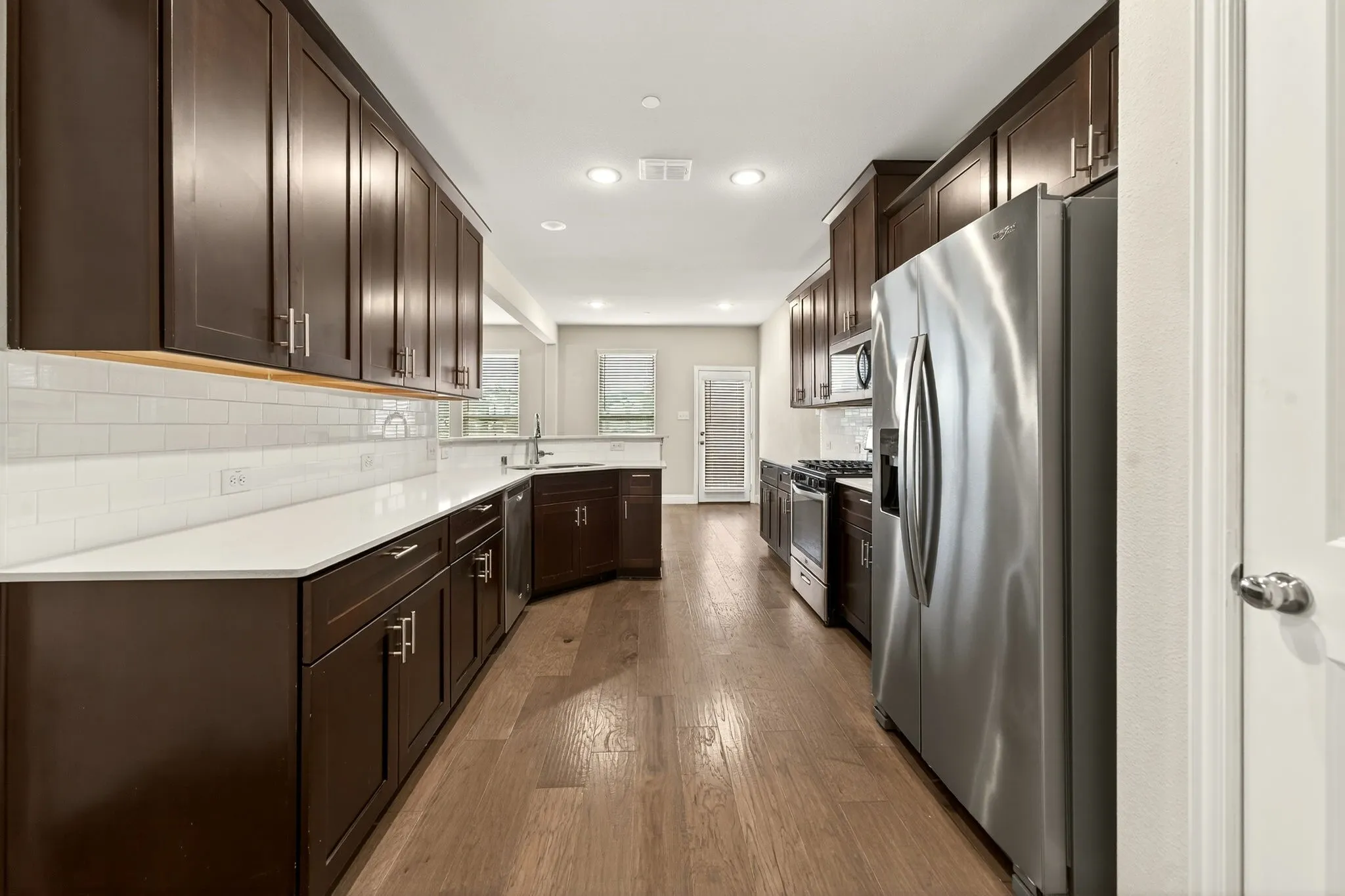Galley kitchen featuring dark wood cabinetry, stainless steel appliances, a white subway tile backsplash, light-toned countertops, and wood-finish flooring