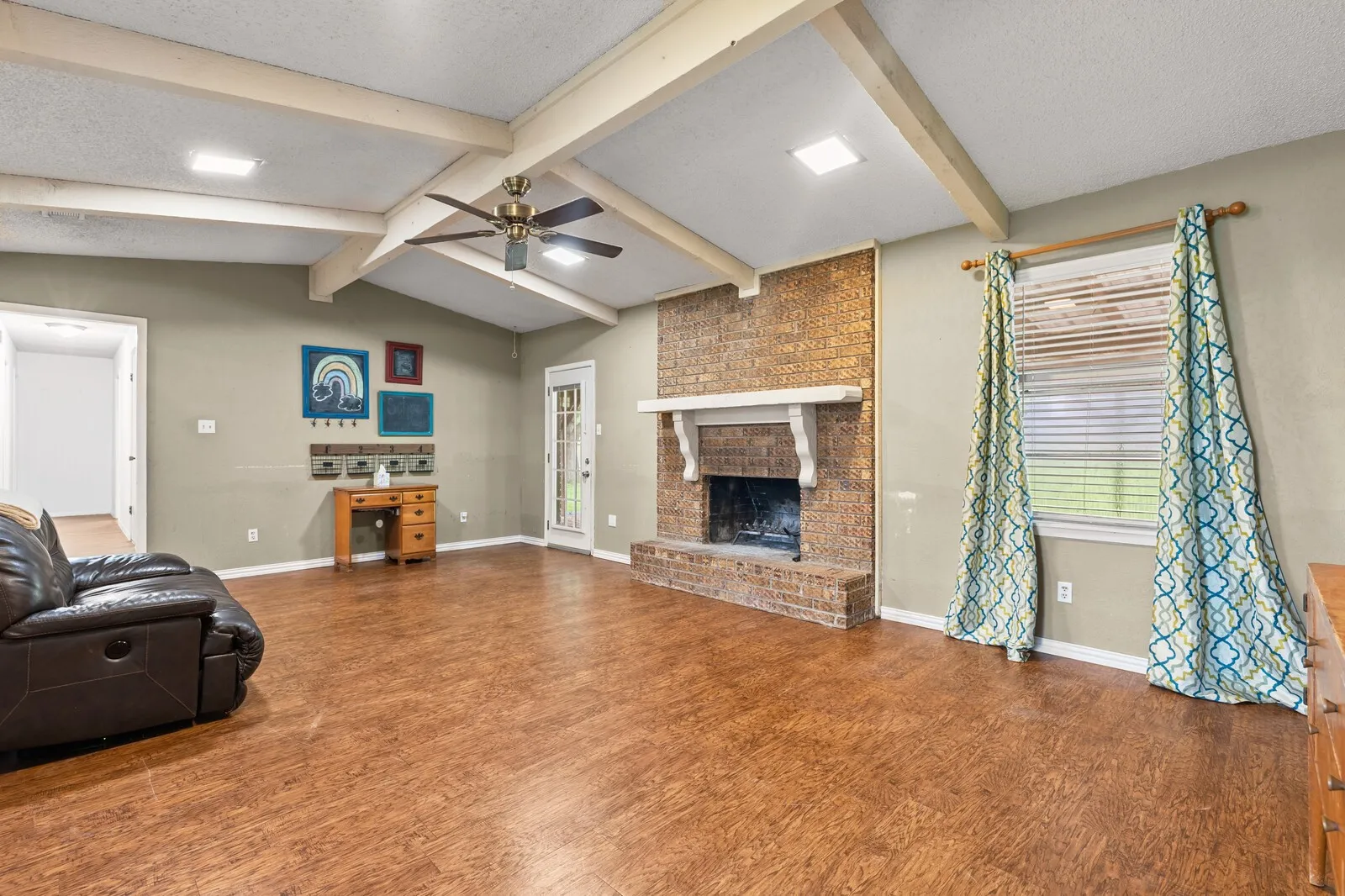 Spacious room featuring vaulted ceilings with exposed beams, wood-finish flooring, and a brick fireplace with a white mantel