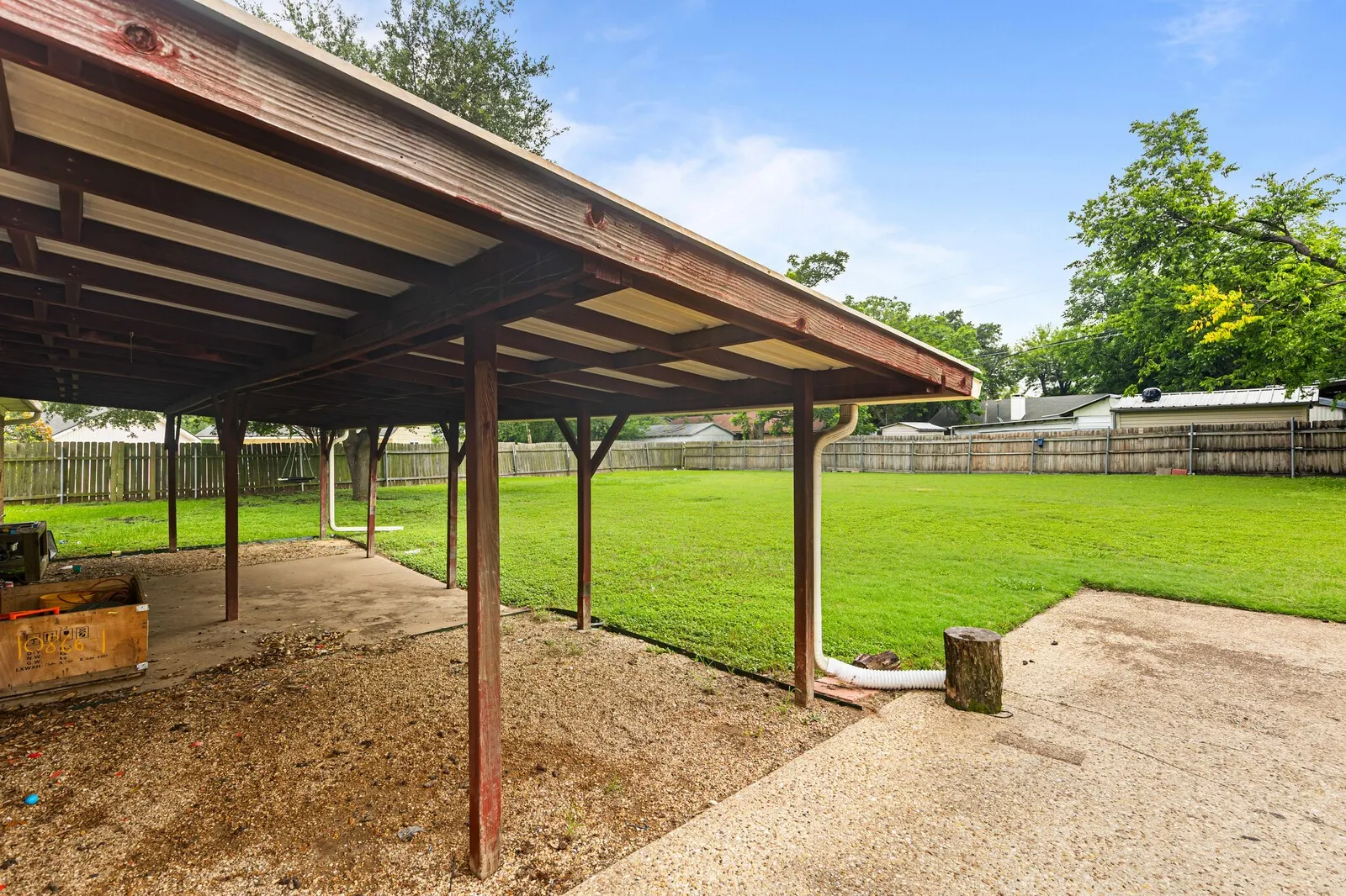 Expansive backyard featuring a large green lawn, perimeter wood fencing, a covered patio area with exposed wood beams, and an additional concrete patio space