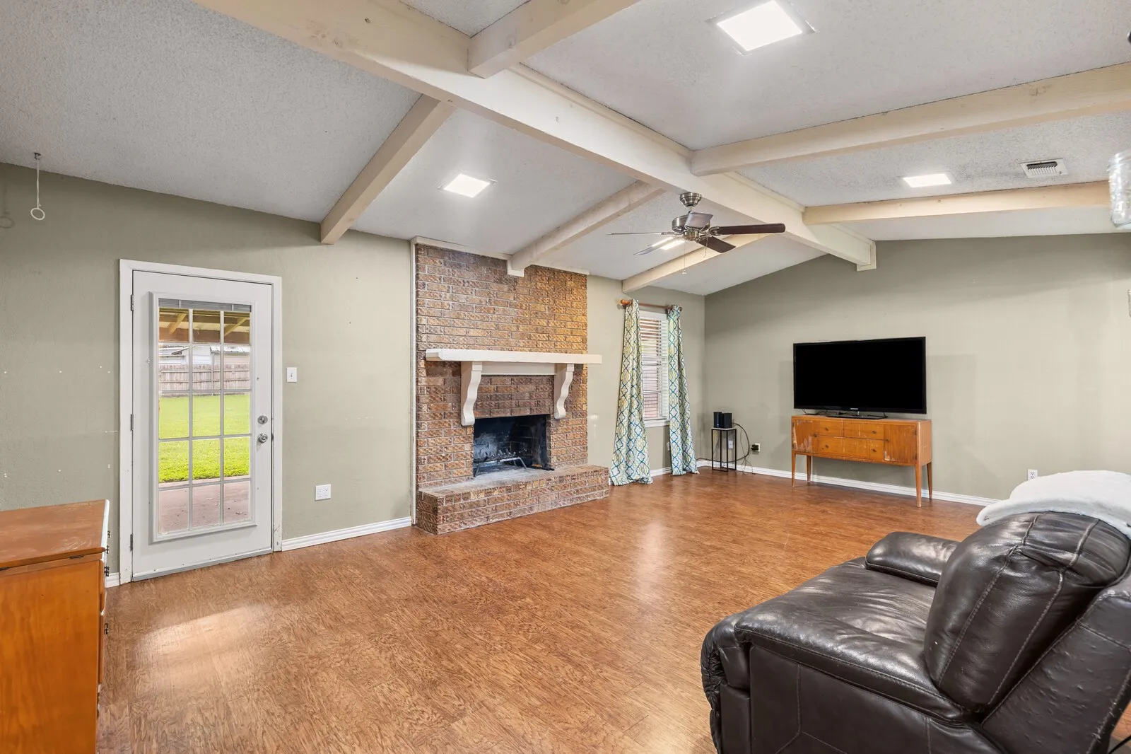 Spacious living area featuring a vaulted ceiling with exposed beams, wood-finish flooring, a brick fireplace with a decorative mantel, and recessed lighting