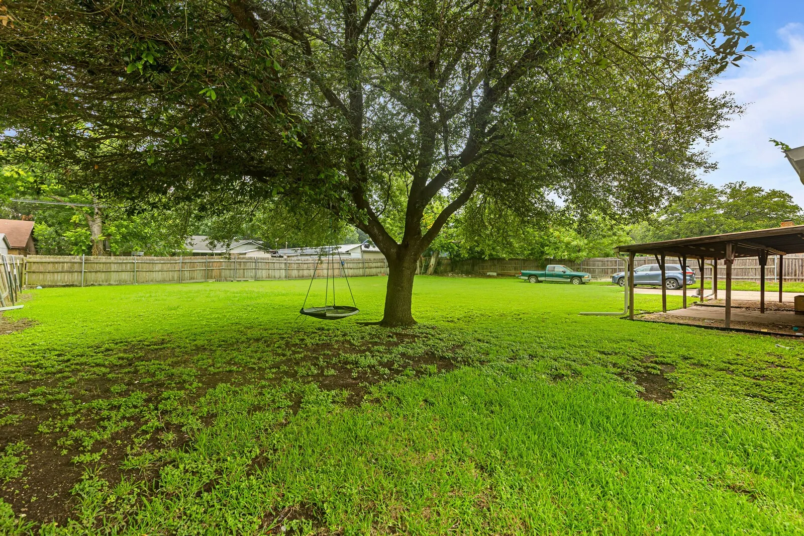Expansive green lawn featuring a mature shade tree, a wooden privacy fence, and a covered patio structure