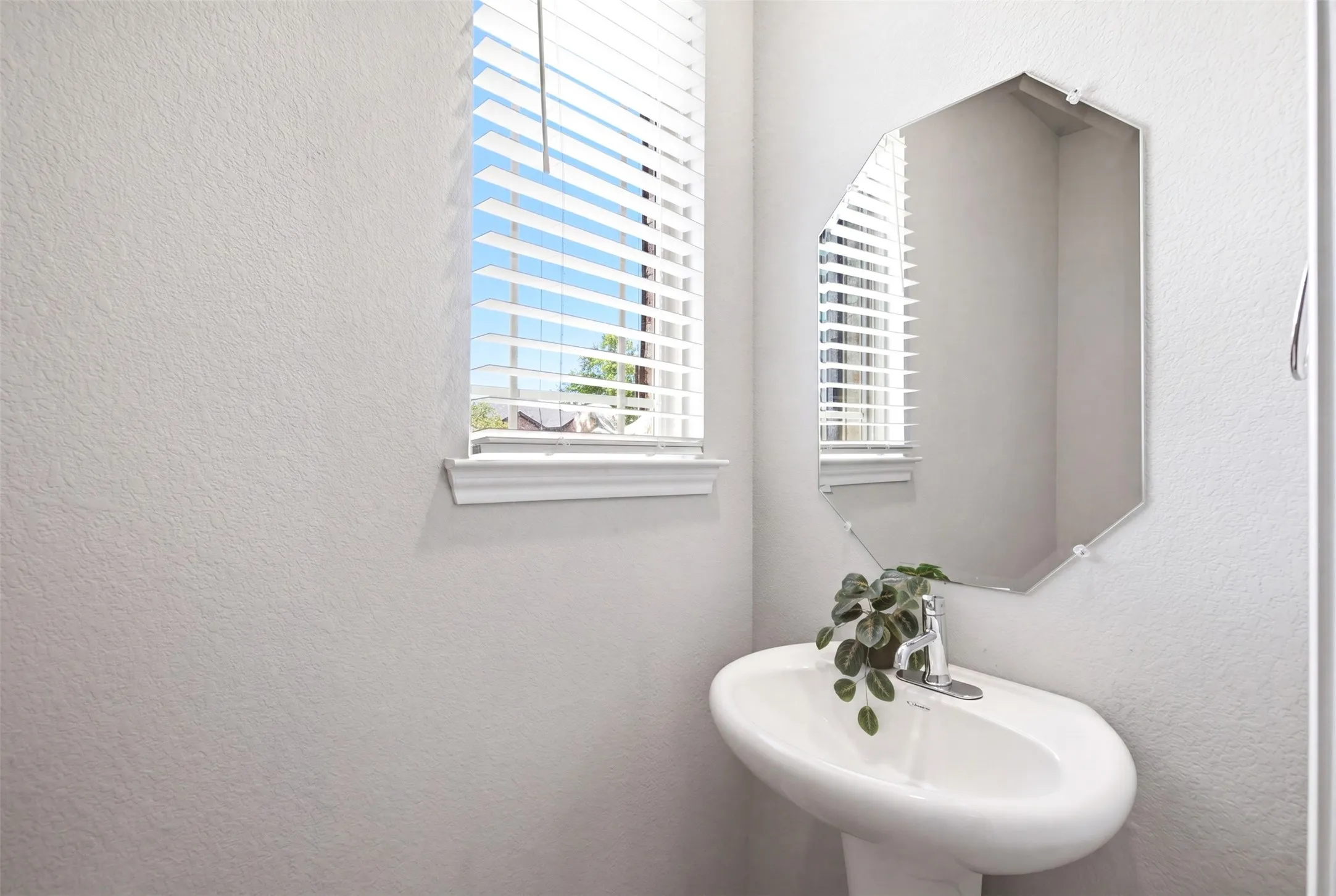 Pedestal sink with a polished chrome faucet, an octagonal wall-mounted mirror, and a window featuring white blinds