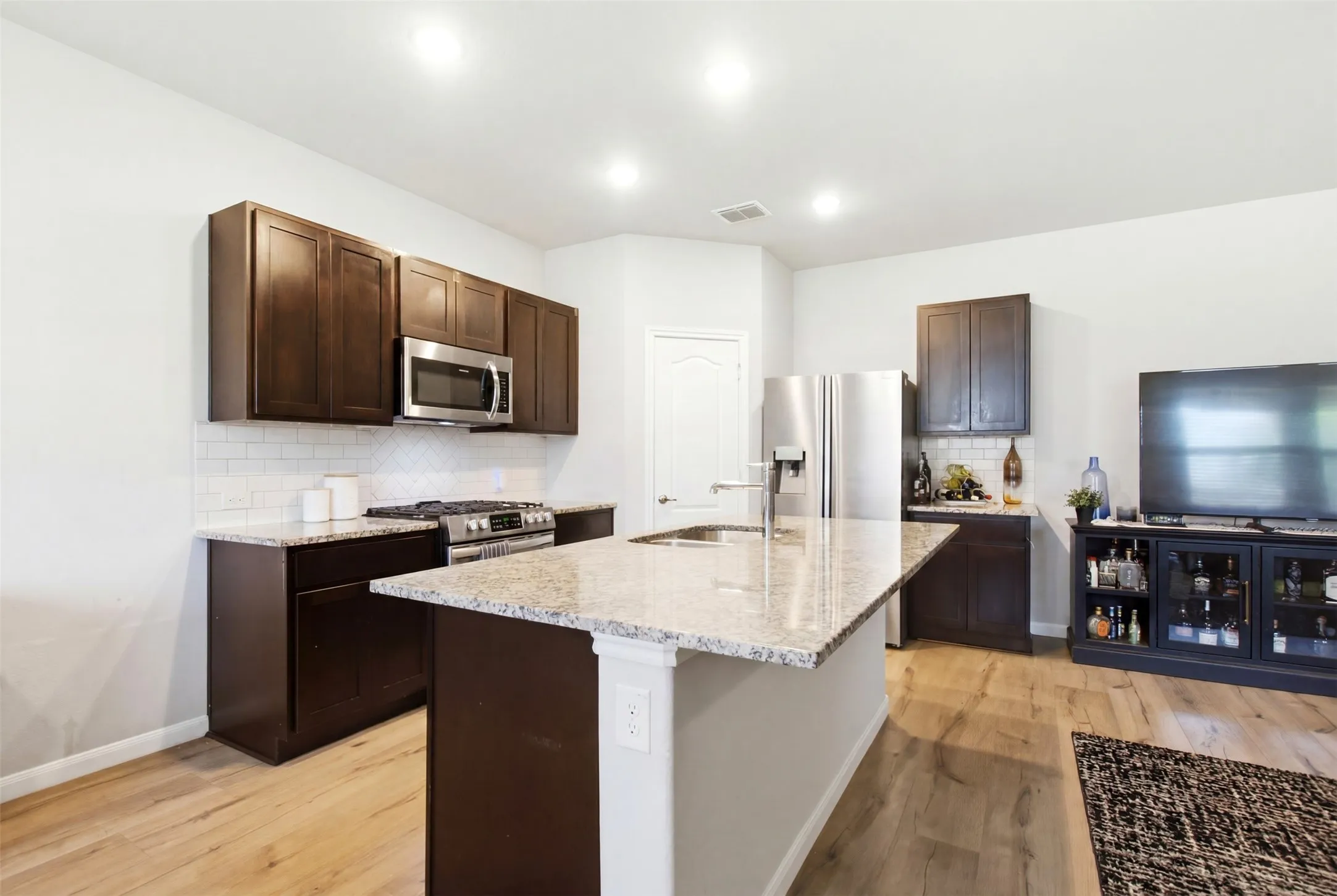 Kitchen featuring dark wood cabinetry, light granite countertops, stainless steel appliances, a subway tile backsplash, and light wood-finish flooring