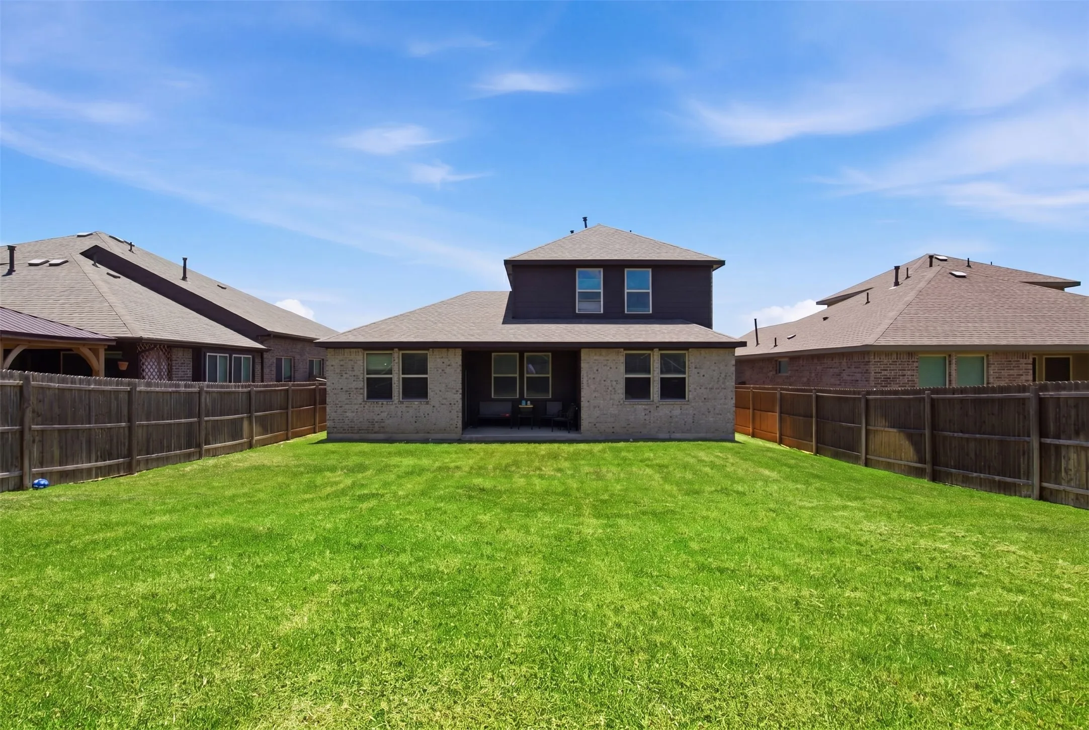 Expansive rear lawn featuring a privacy fence, brick exterior with dark siding, multiple windows, and a covered patio area