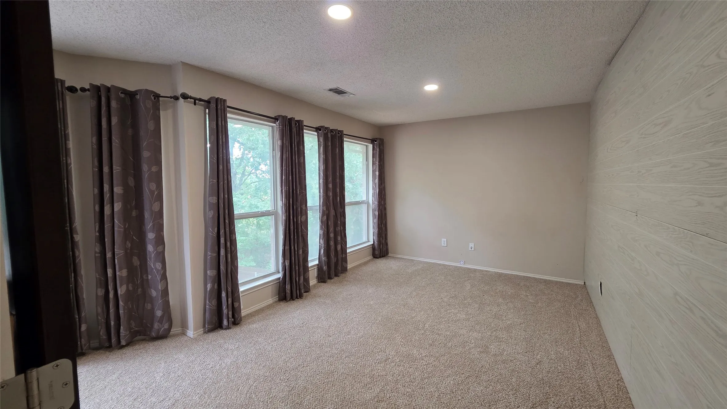 Spacious room with four large windows, neutral carpeting, light-toned walls, recessed lighting, and a prominent wood-finish accent wall
