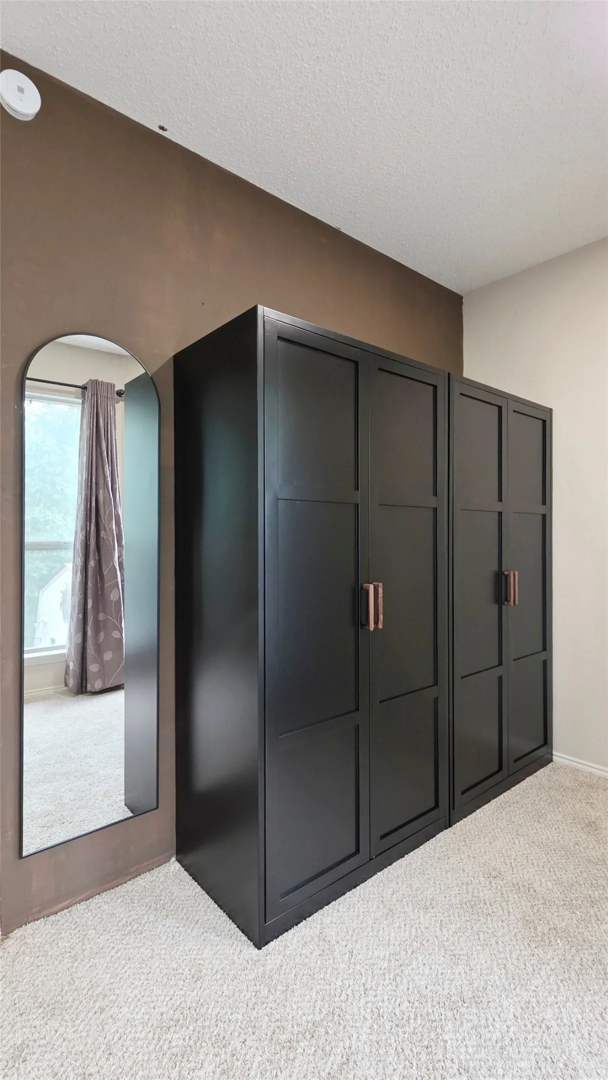 Built-in black cabinetry with bronze hardware, light-toned carpet flooring, a full-length arched mirror, and a window with patterned drapes