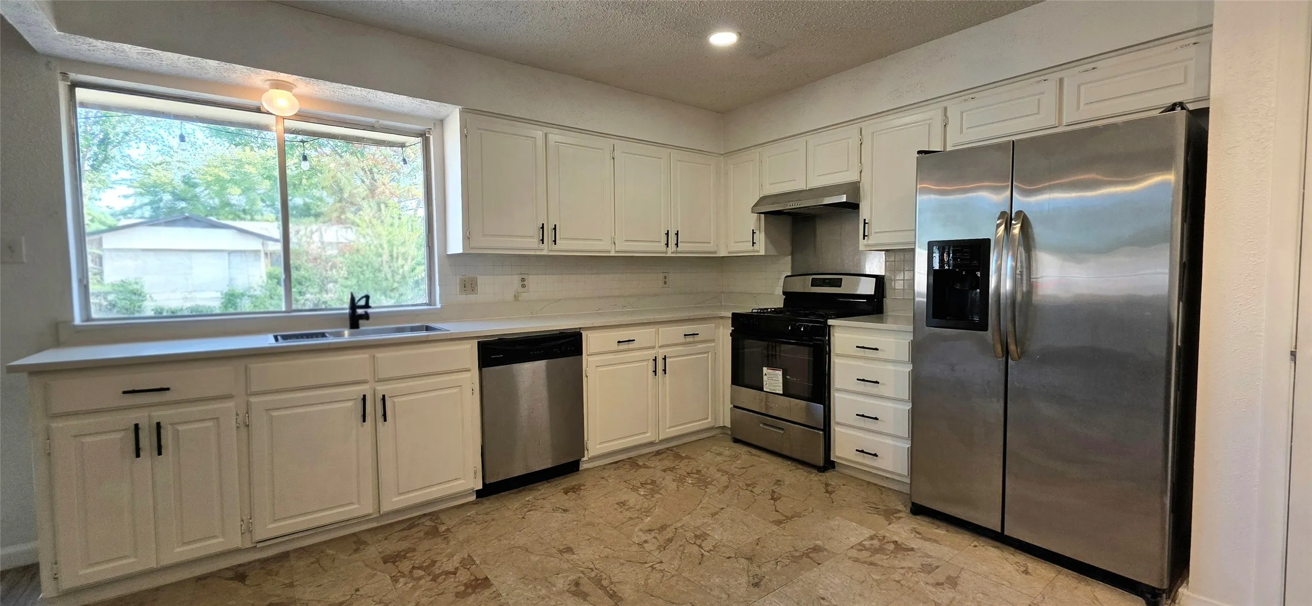 White cabinetry, stainless steel appliances, stone-look flooring, large window, and recessed lighting
