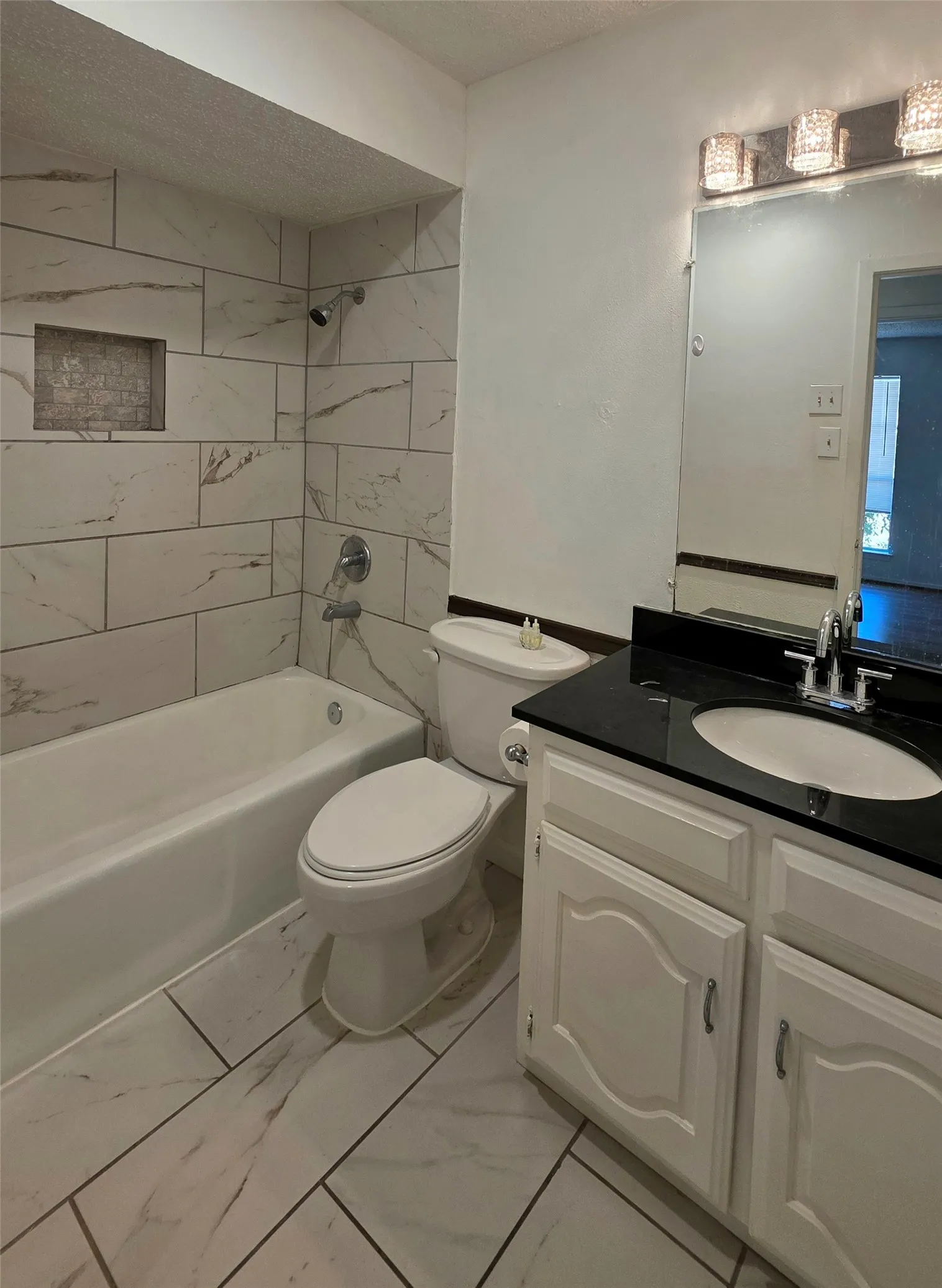 Bathroom featuring a white bathtub, tiled shower surround with a recessed niche, white vanity with a dark countertop, and a vanity mirror with overhead lighting