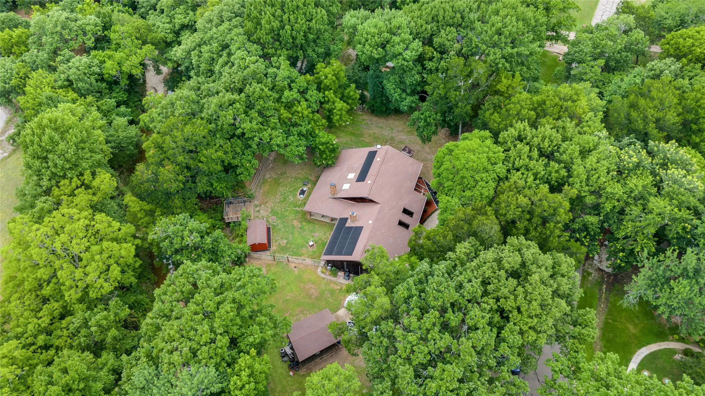 Tree-surrounded property featuring a main residence with a brown shingle roof, multiple solar panels, and an attached deck