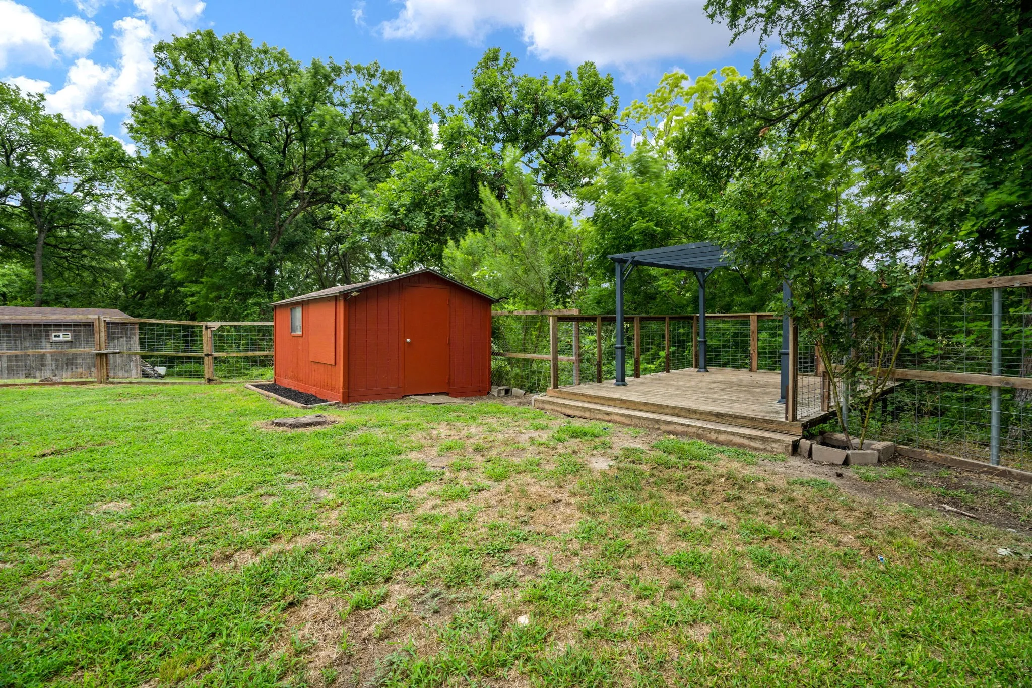 Expansive outdoor space featuring a raised wooden deck with a dark metal pergola, a red storage shed, and a wooden fence with wire mesh