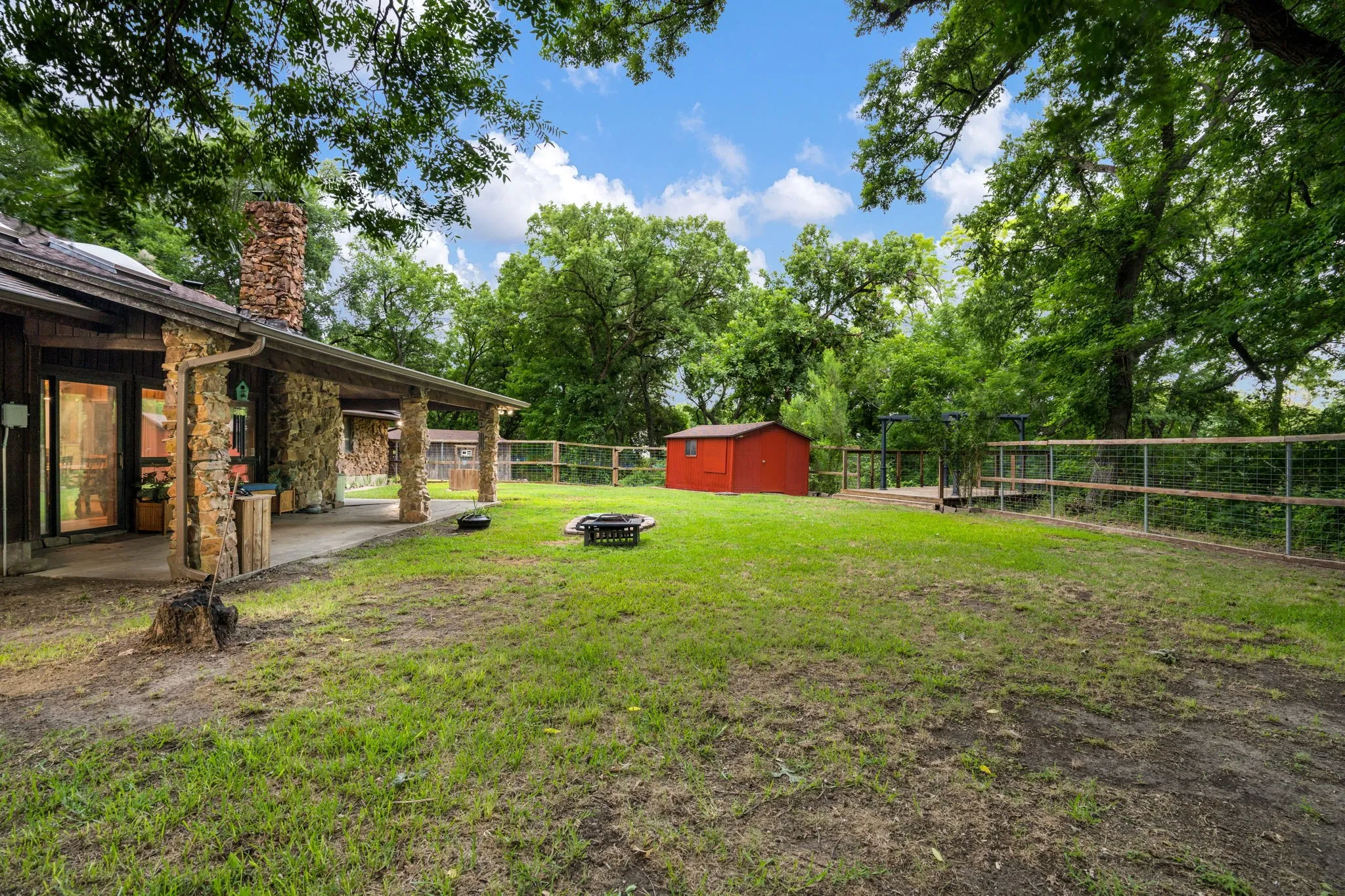 Expansive green space featuring a stone-clad exterior, stone chimney, covered patio, a red utility shed, and perimeter fencing