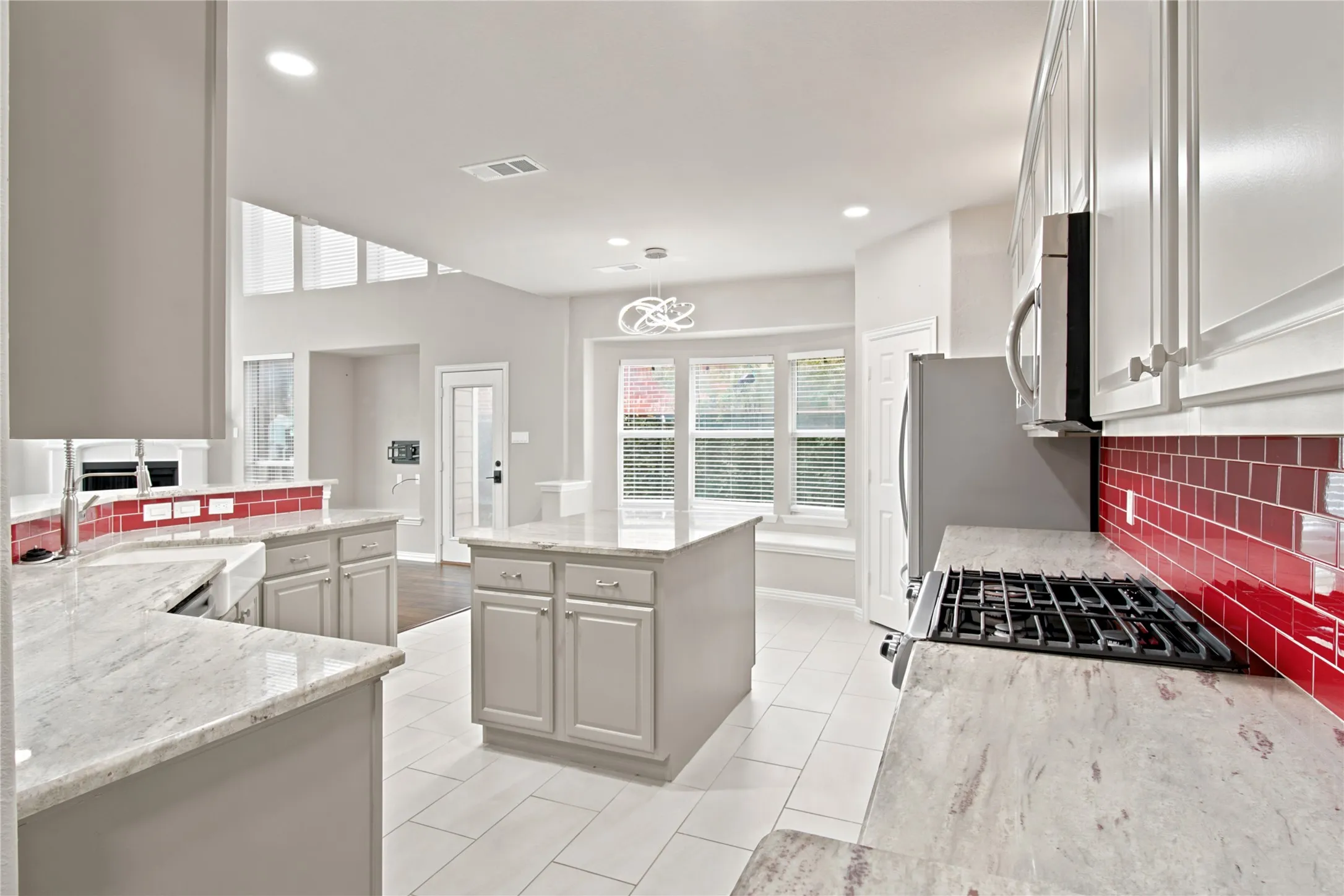 Spacious kitchen featuring an island, light-toned countertops, contrasting red tile backsplash, stainless steel appliances, and white tile flooring