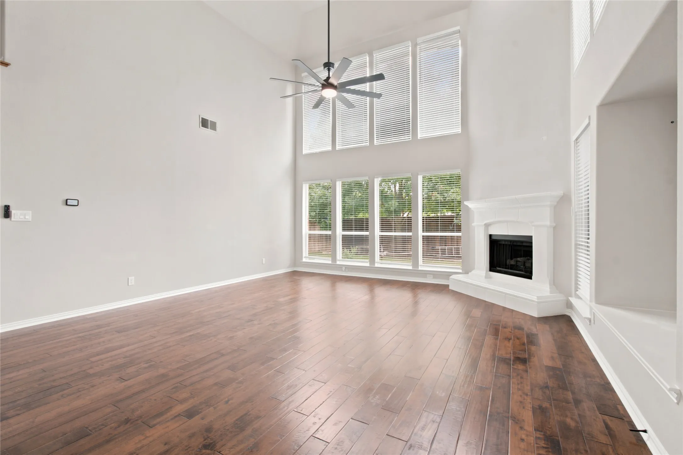 Spacious living area featuring high ceilings, wood-finish flooring, a corner fireplace with white mantel, a ceiling fan, and tall windows providing abundant natural light