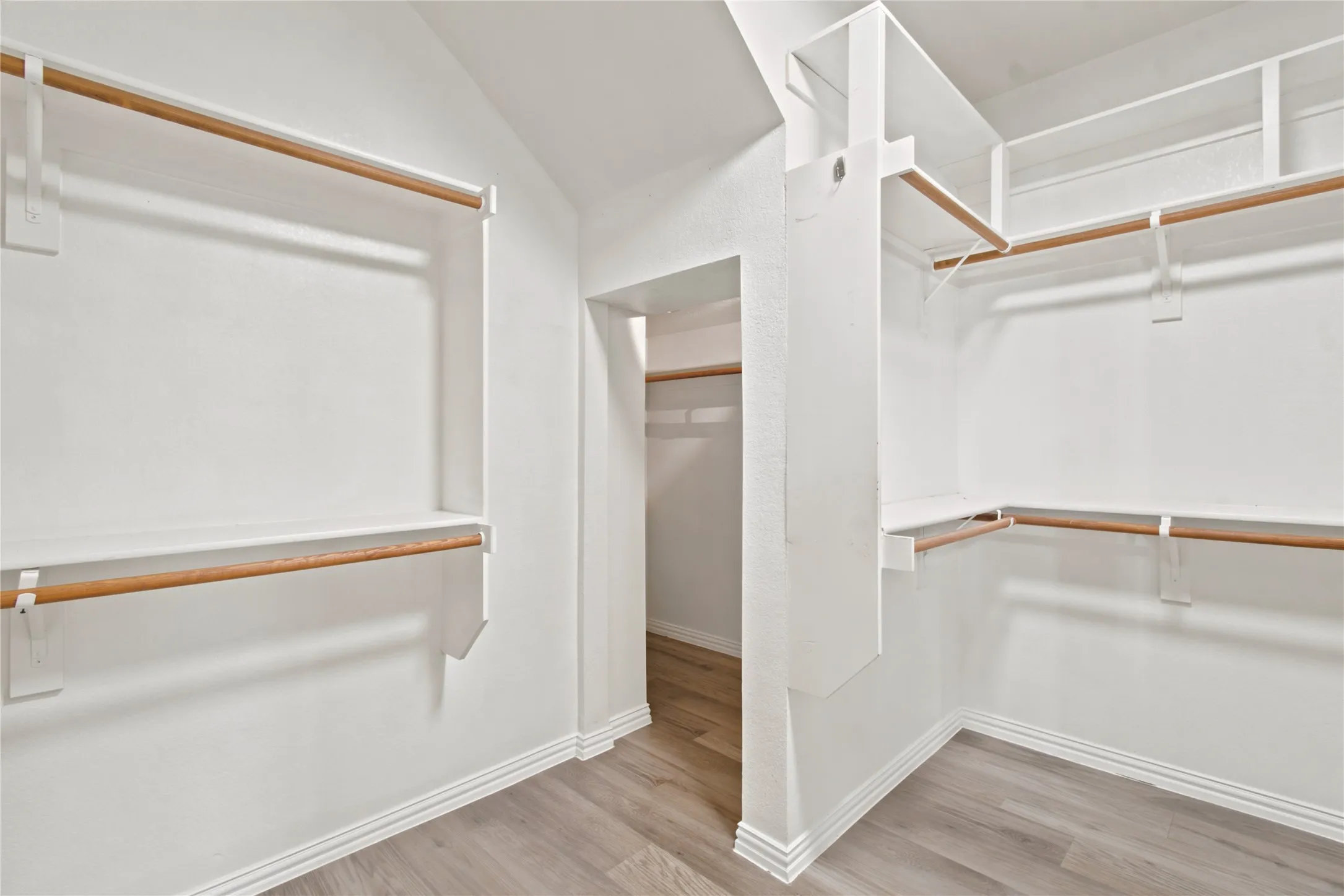 Walk-in closet featuring light wood-finish flooring, white baseboards, multiple white shelving units, and natural wood hanging rods