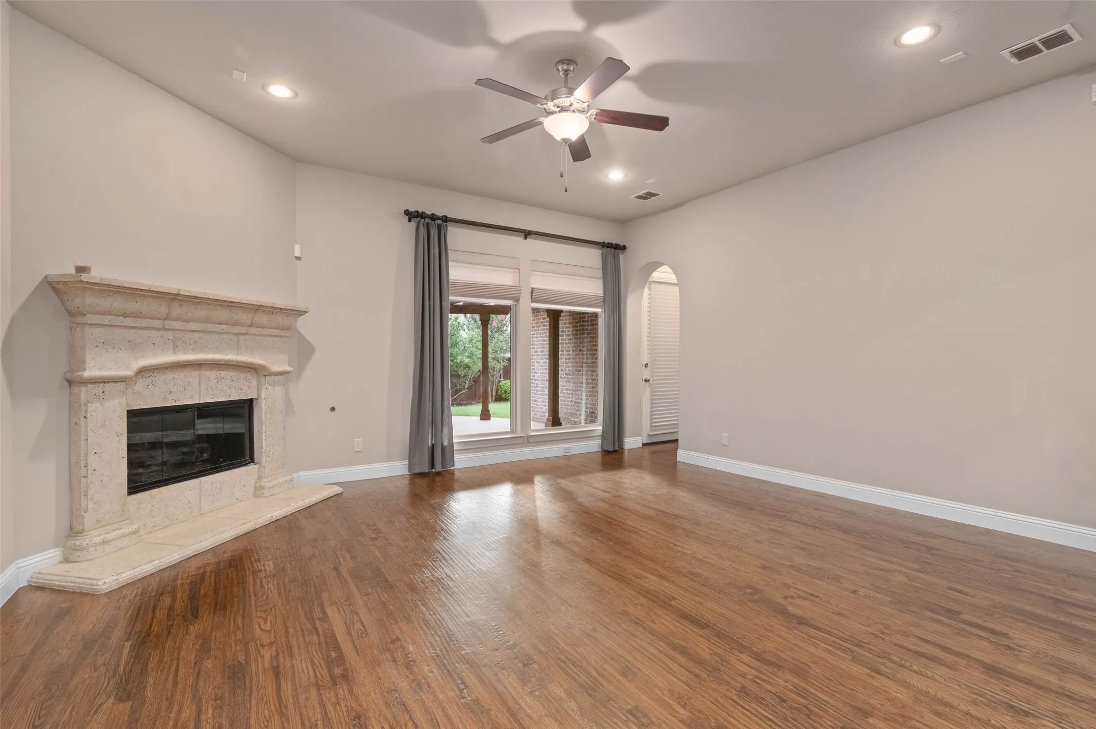Spacious living area featuring rock fireplace, ceiling fan, wood flooring and large windows allowing in plenty of natural light.