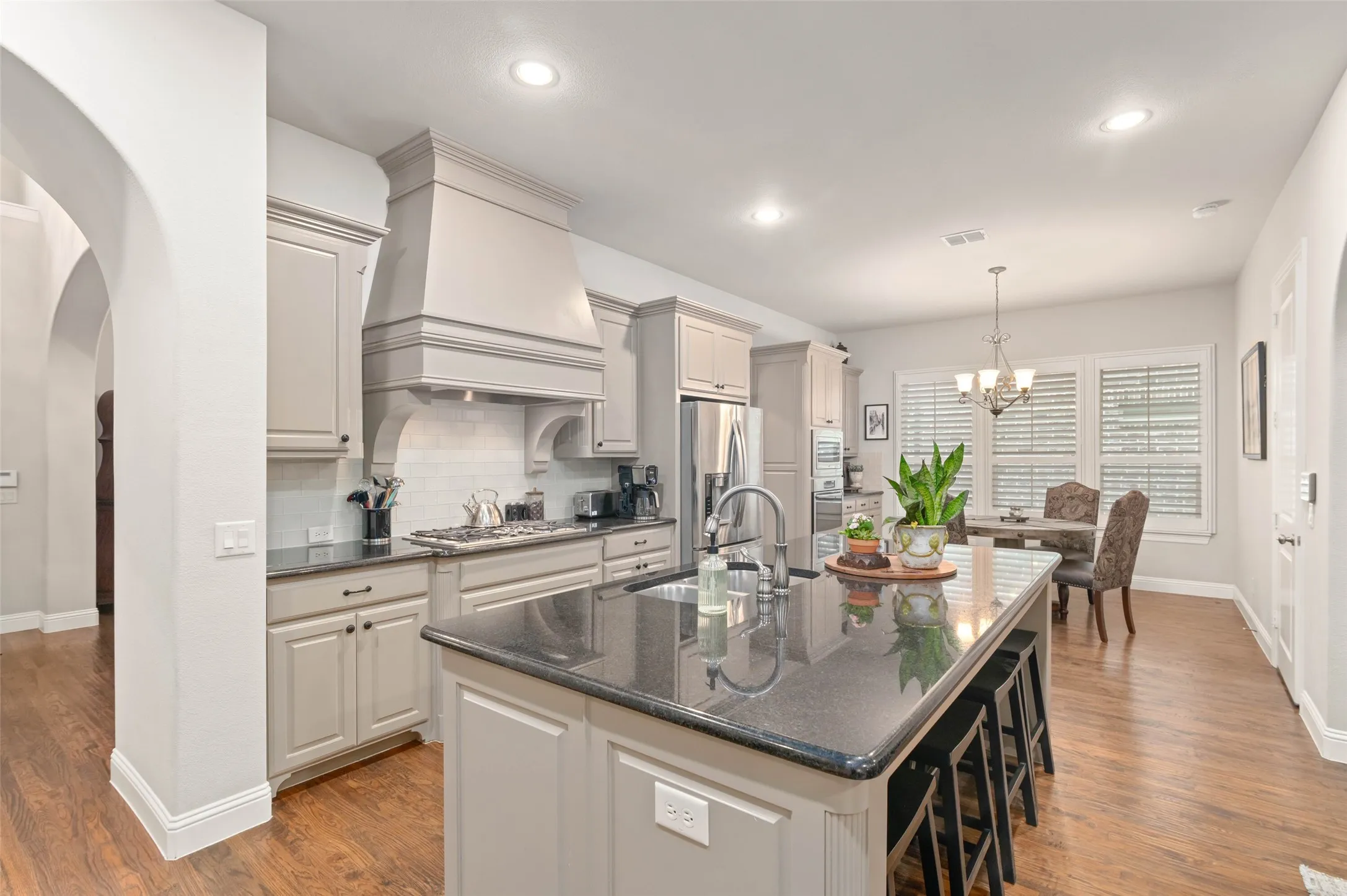 Open-concept kitchen featuring an oversized island with seating, dark stone countertops, wood-finish flooring, recessed lighting, and a dining area with plantation shutters