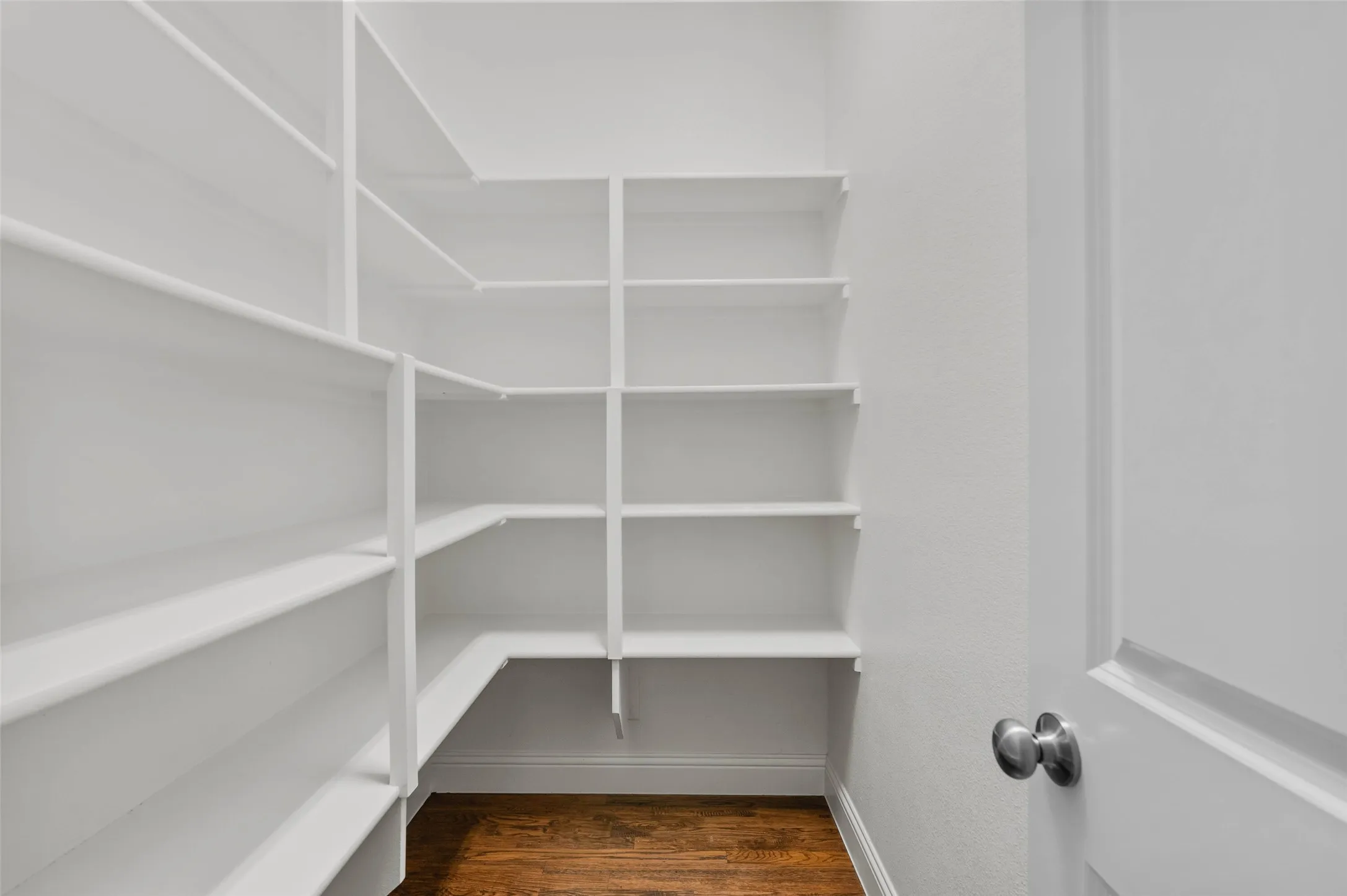 Spacious walk-in pantry featuring white shelving, hardwood flooring, and a panel door with a satin nickel knob