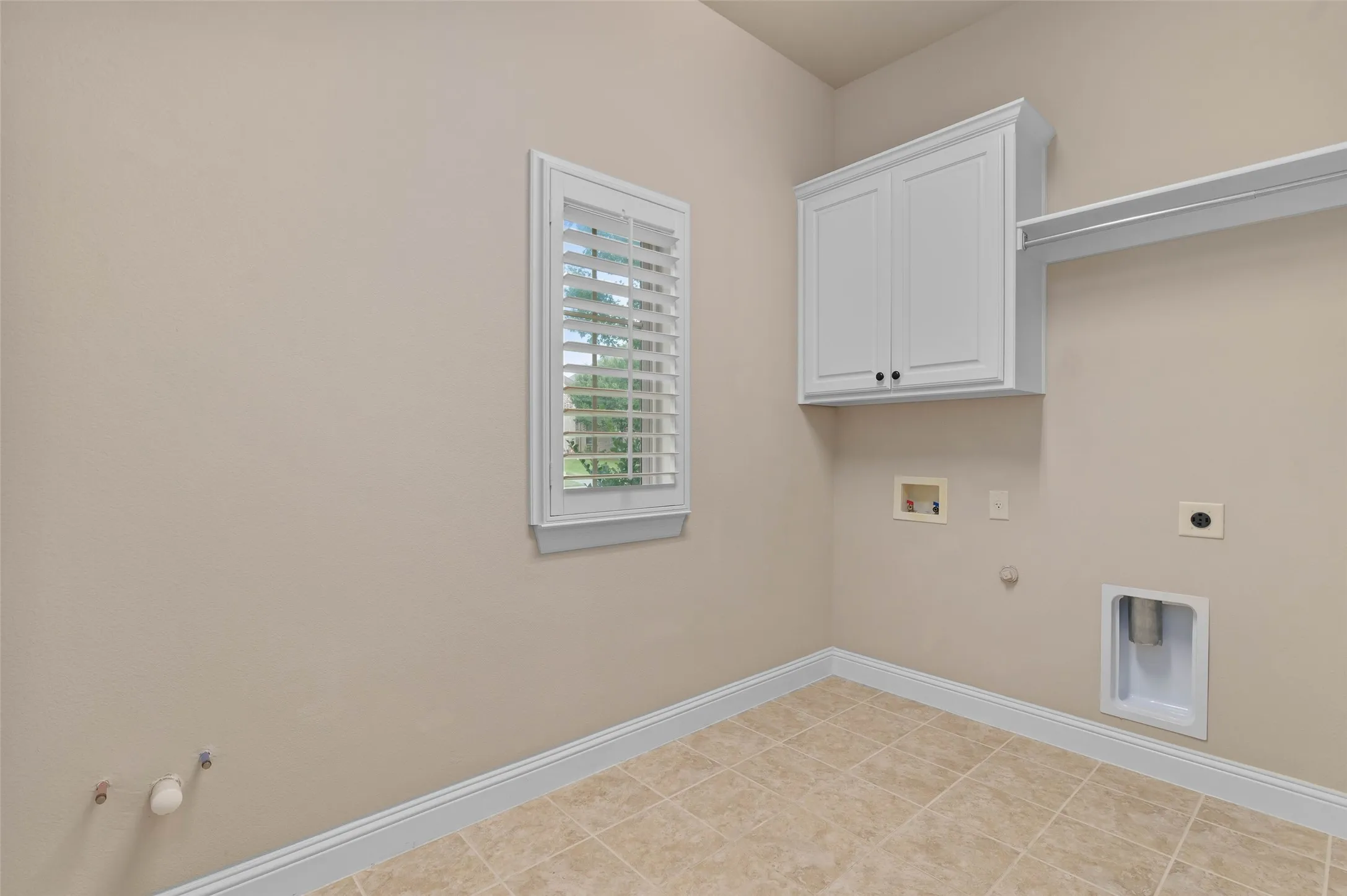 Dedicated utility space featuring a window with plantation shutters, tile flooring, white cabinetry, and a built-in shelf