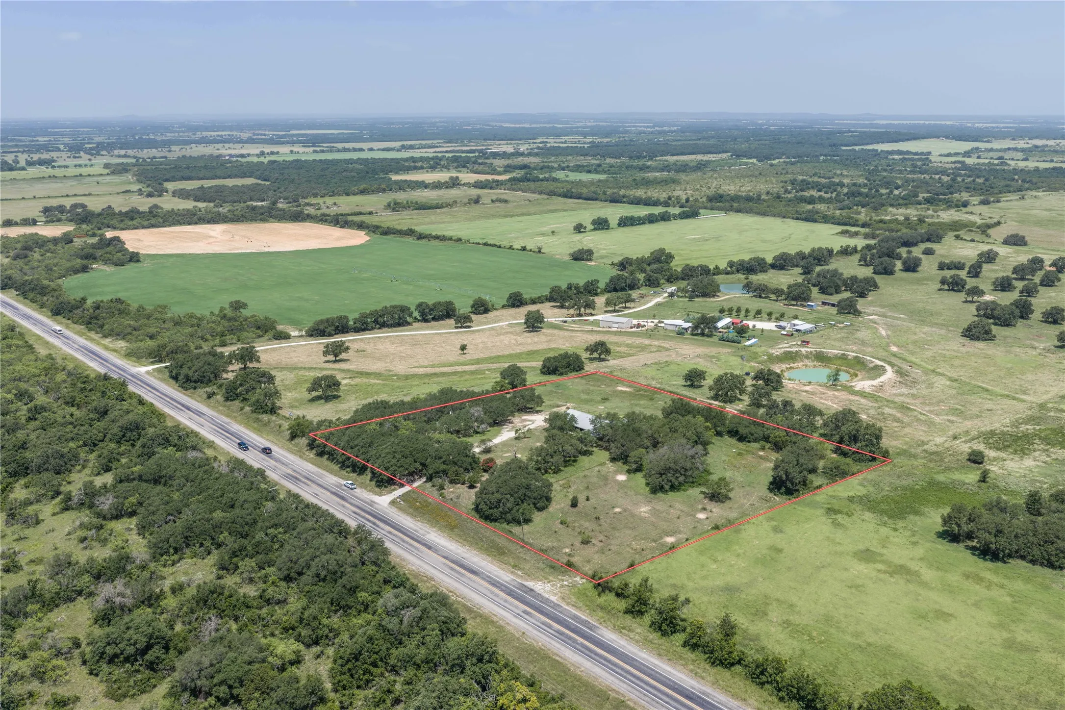 Expansive undeveloped land featuring a perimeter fence, mature trees, open grassy areas, and direct road frontage