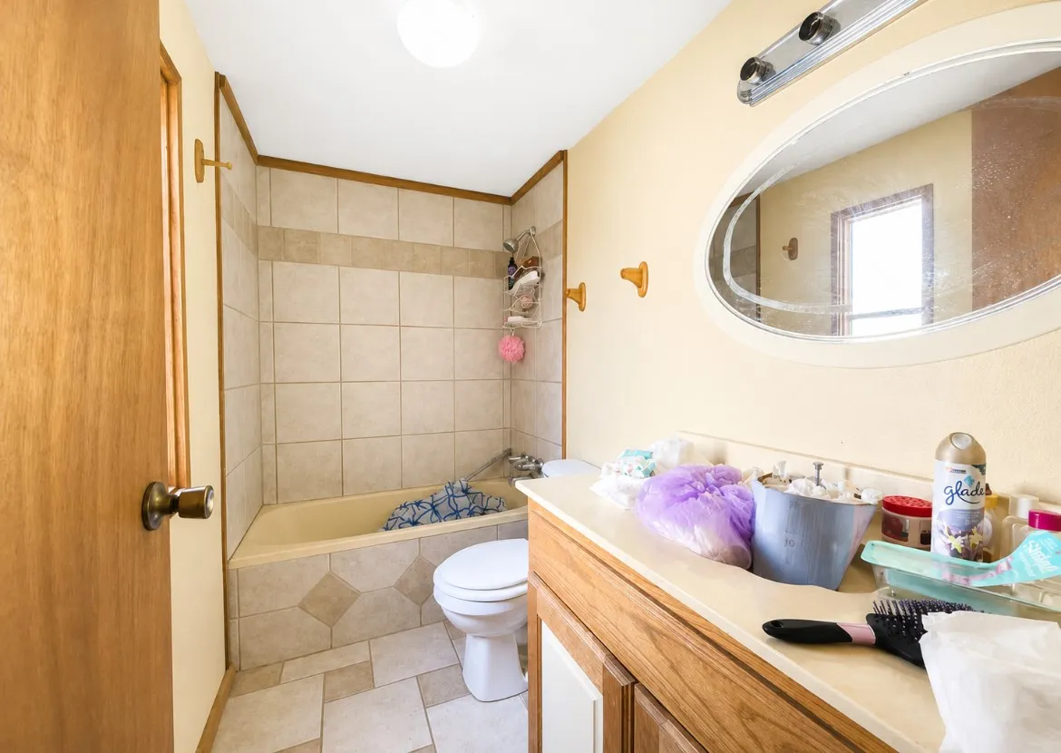 Bathroom featuring a built-in tub with tile surround, a single vanity with laminate countertop, and an oval mirror with overhead bar​​‌​​​​‌​​‌‌​‌‌​​​‌‌​‌​‌​‌​​​‌​​ lighting