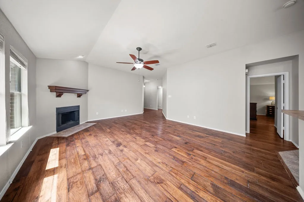Spacious living area featuring wood-finish flooring, a corner fireplace with a wooden mantel, and a ceiling fan