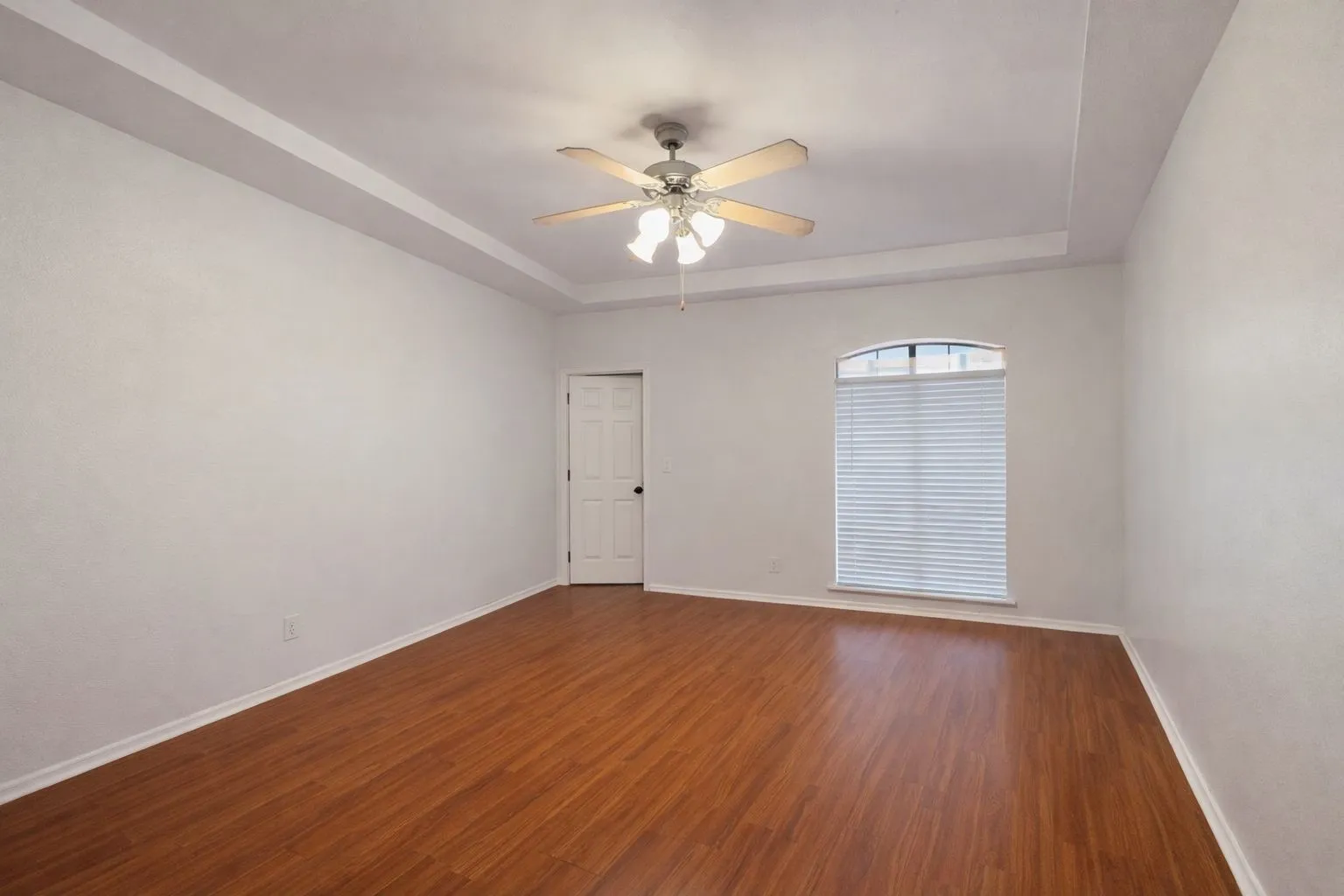Spacious room featuring wood-finish flooring, a tray ceiling with a fan light fixture, an arched window with blinds, and white baseboards