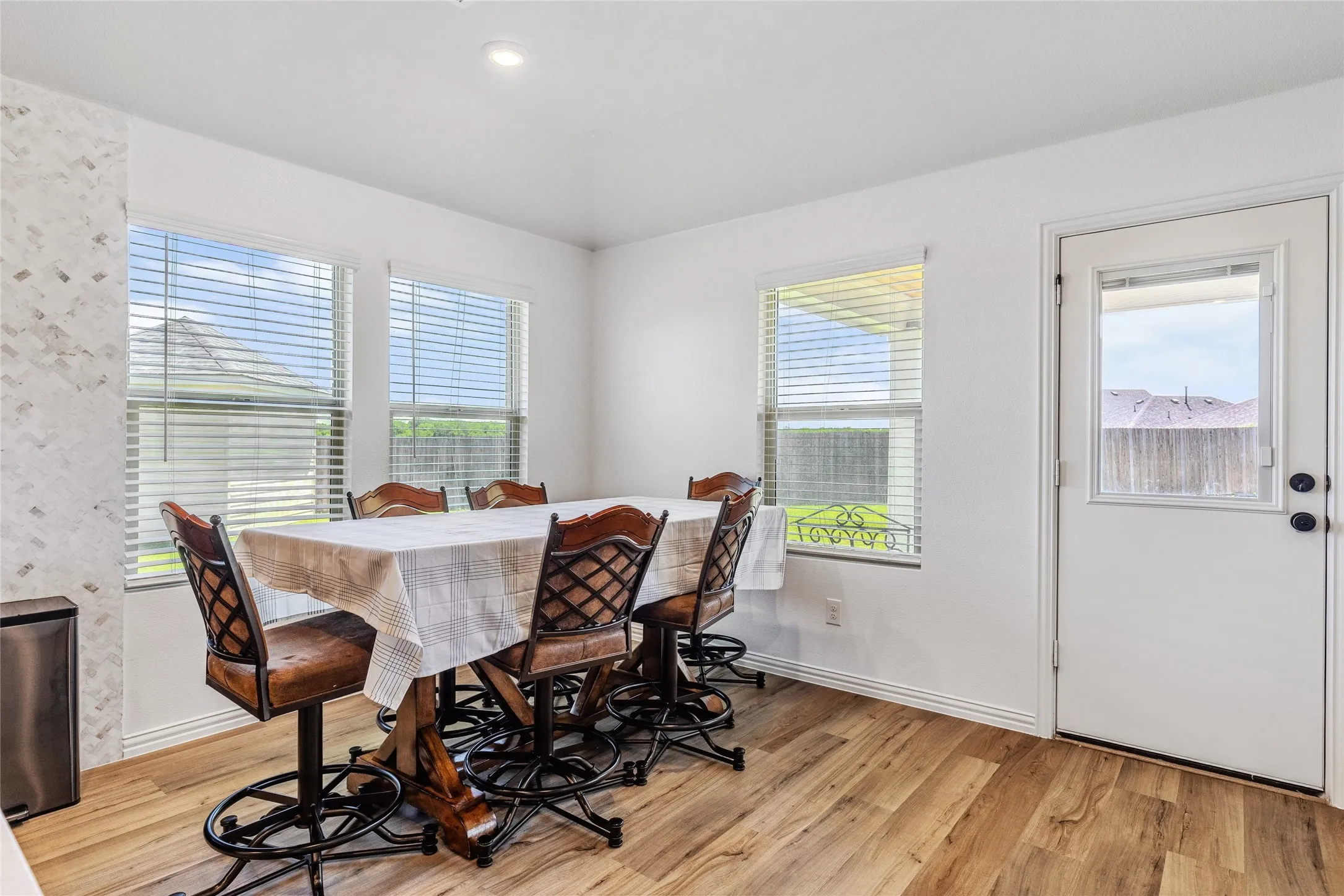 Dining area featuring wood-finish flooring, two window openings with blinds, a side door with integrated window, white baseboards, and a decorative tile accent wall
