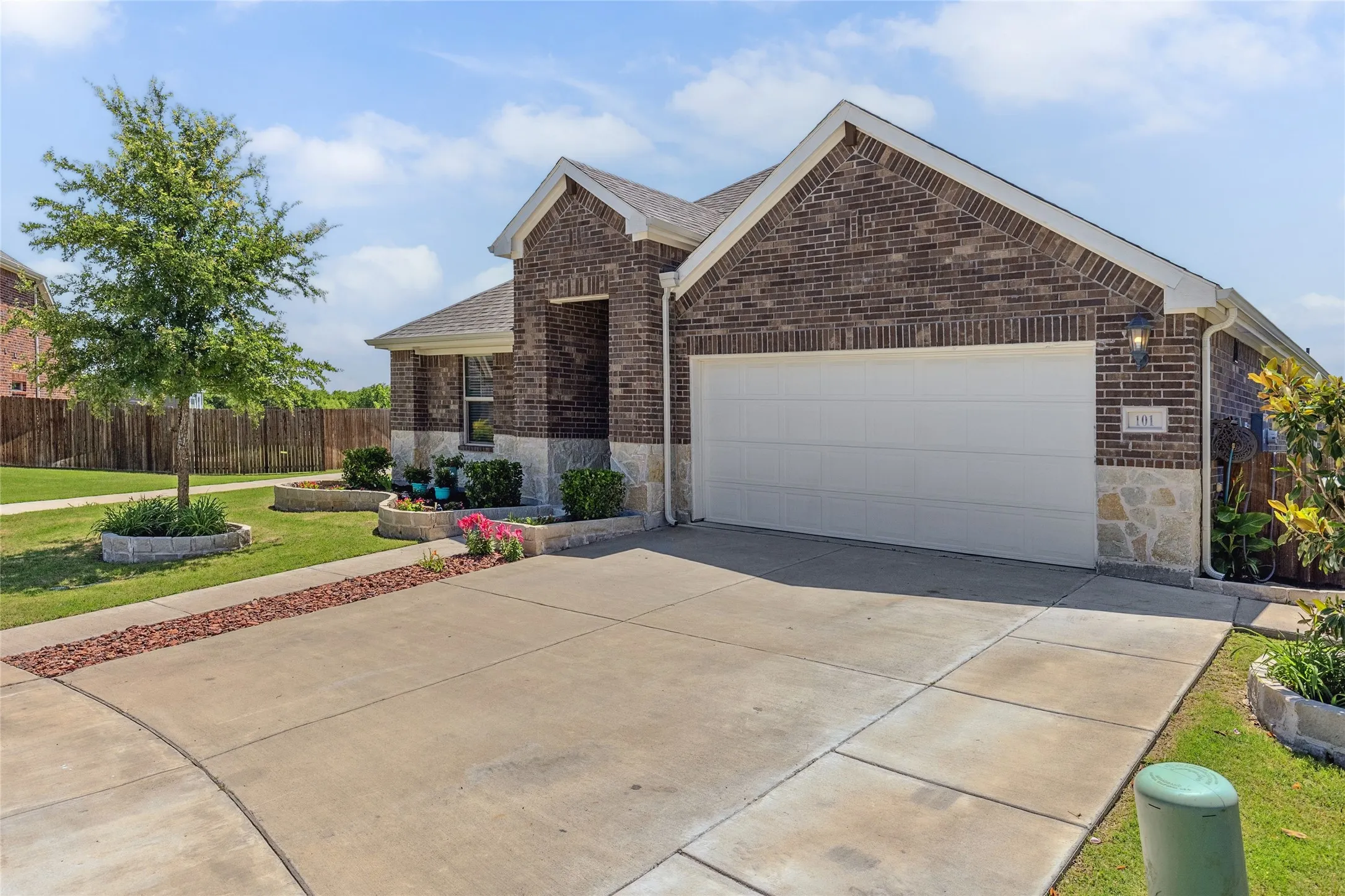 Brick and stone facade featuring a two-car garage, concrete driveway, and landscaped front yard with a mature tree and garden beds