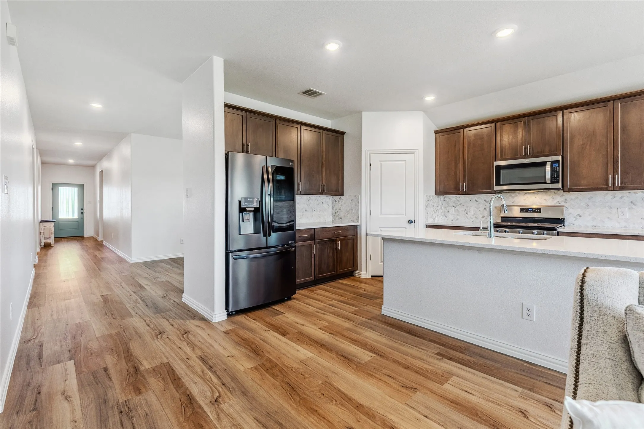 Open-concept kitchen featuring wood-finish flooring, dark wood cabinetry, stainless steel appliances, a white tile backsplash, and a white countertop island with an integrated sink