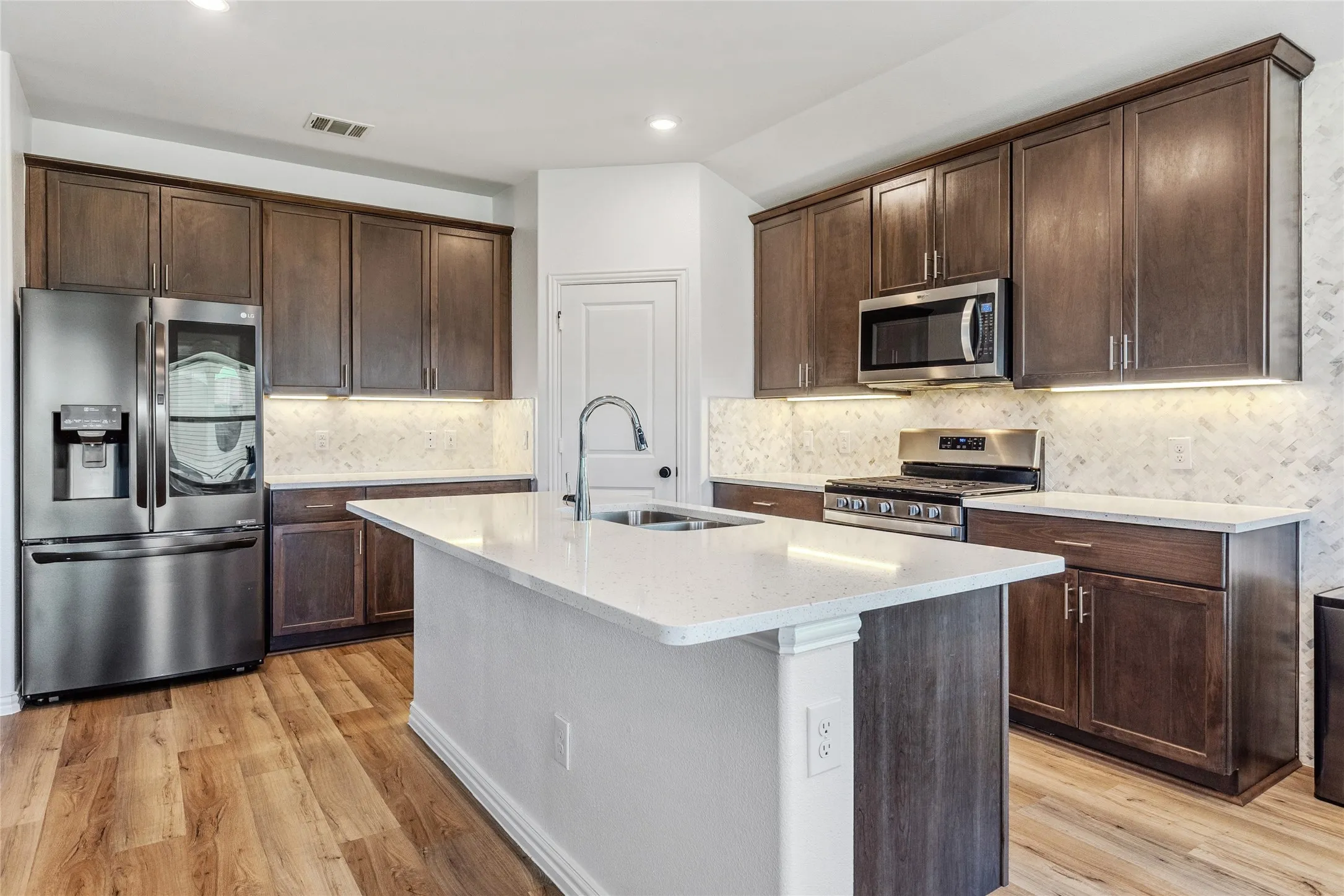 Kitchen featuring a central island with an undermount sink, ample dark wood-finish cabinetry, light-toned countertops, a patterned tile backsplash, and stainless steel appliances including a refrigerator, microwave, and gas range