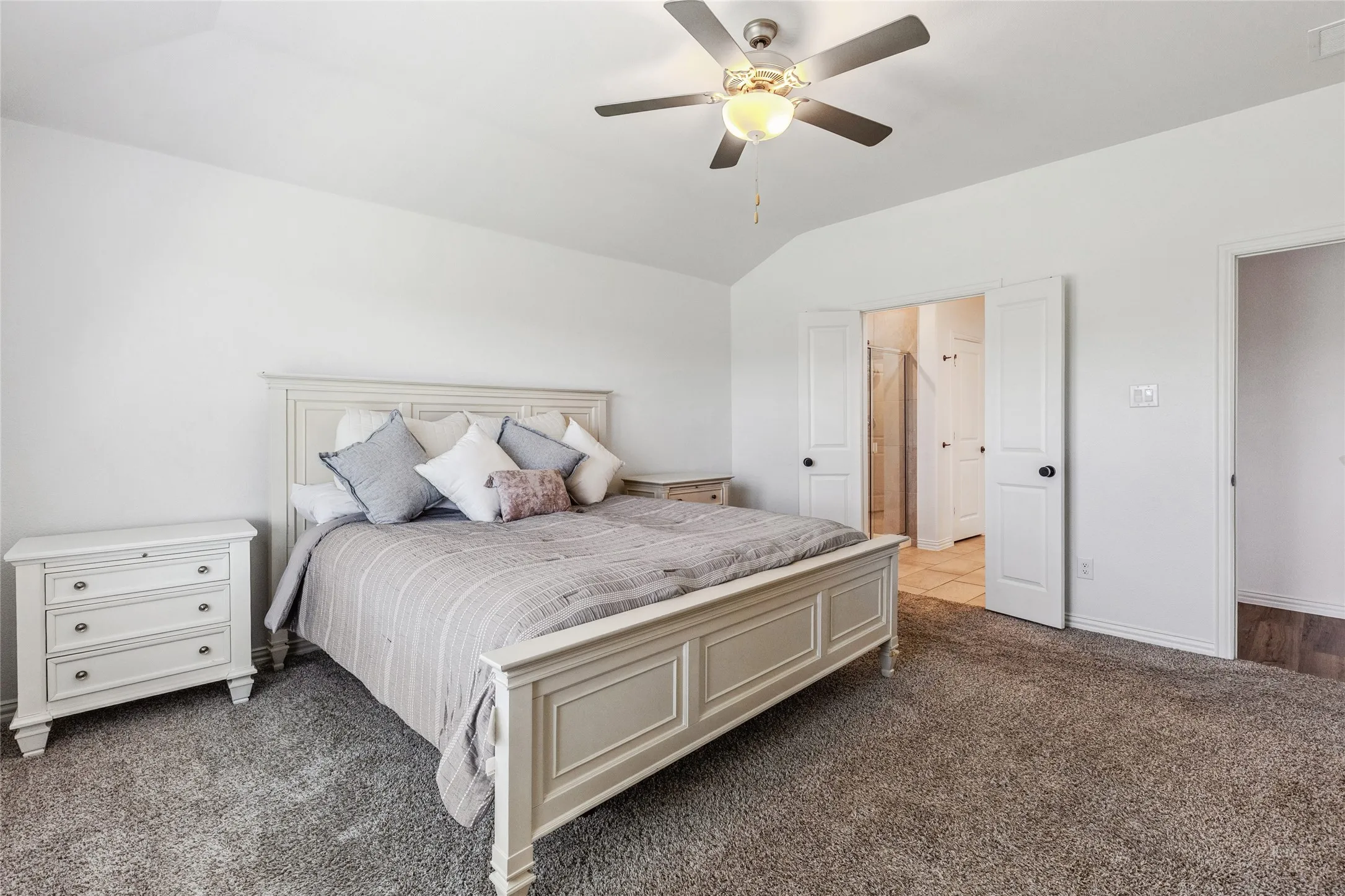 Carpeted bedroom featuring a vaulted ceiling with a ceiling fan, white walls, and double doors opening to a tiled bathroom with a glass-enclosed shower