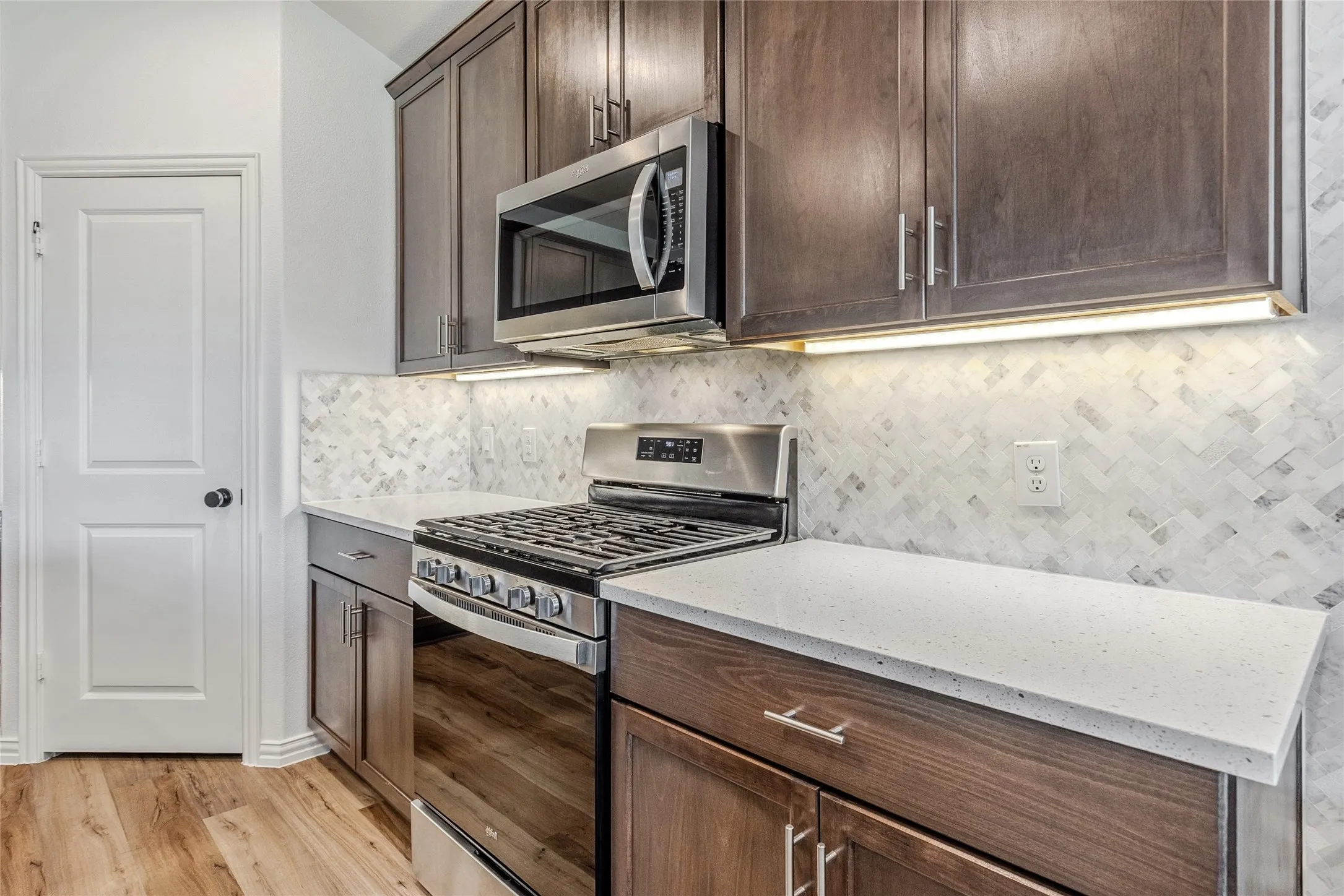 Kitchen featuring stainless steel appliances, wood-finish cabinetry, light-toned countertops, tile backsplash, and wood-finish flooring