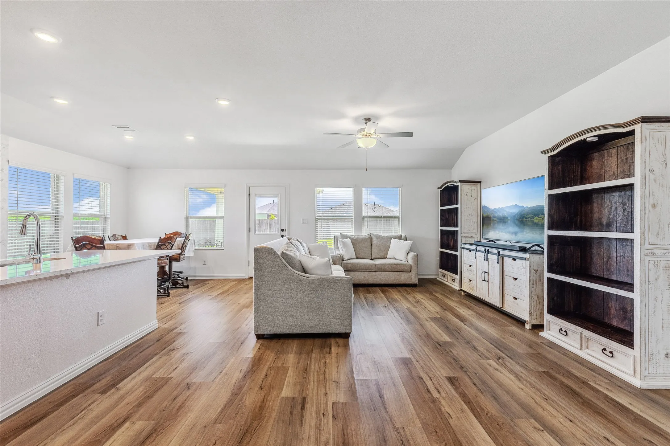 Expansive open-concept living area featuring wood-finish flooring, vaulted ceilings, recessed lighting, and multiple windows with blinds